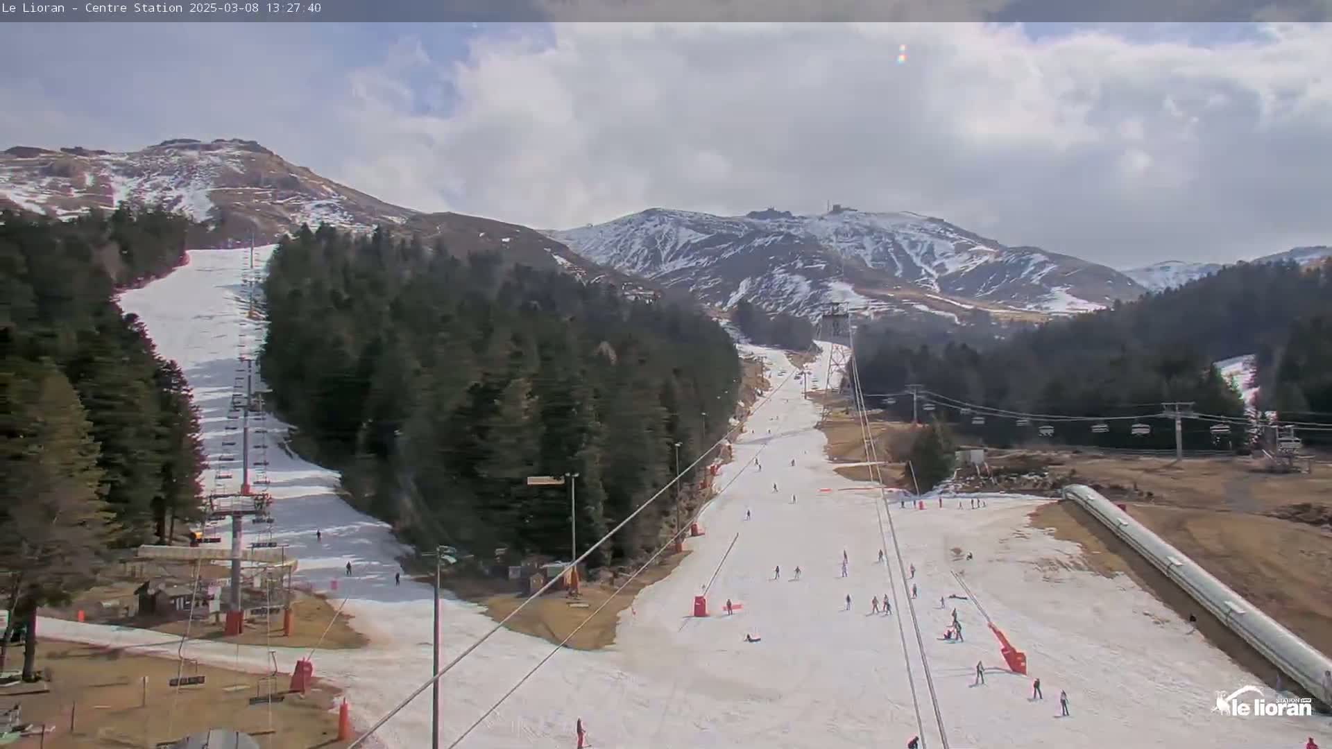 A mostly sunny day reveals a ski slope with several skiers and snowboarders, chairlifts, and snow-covered mountains in the background.