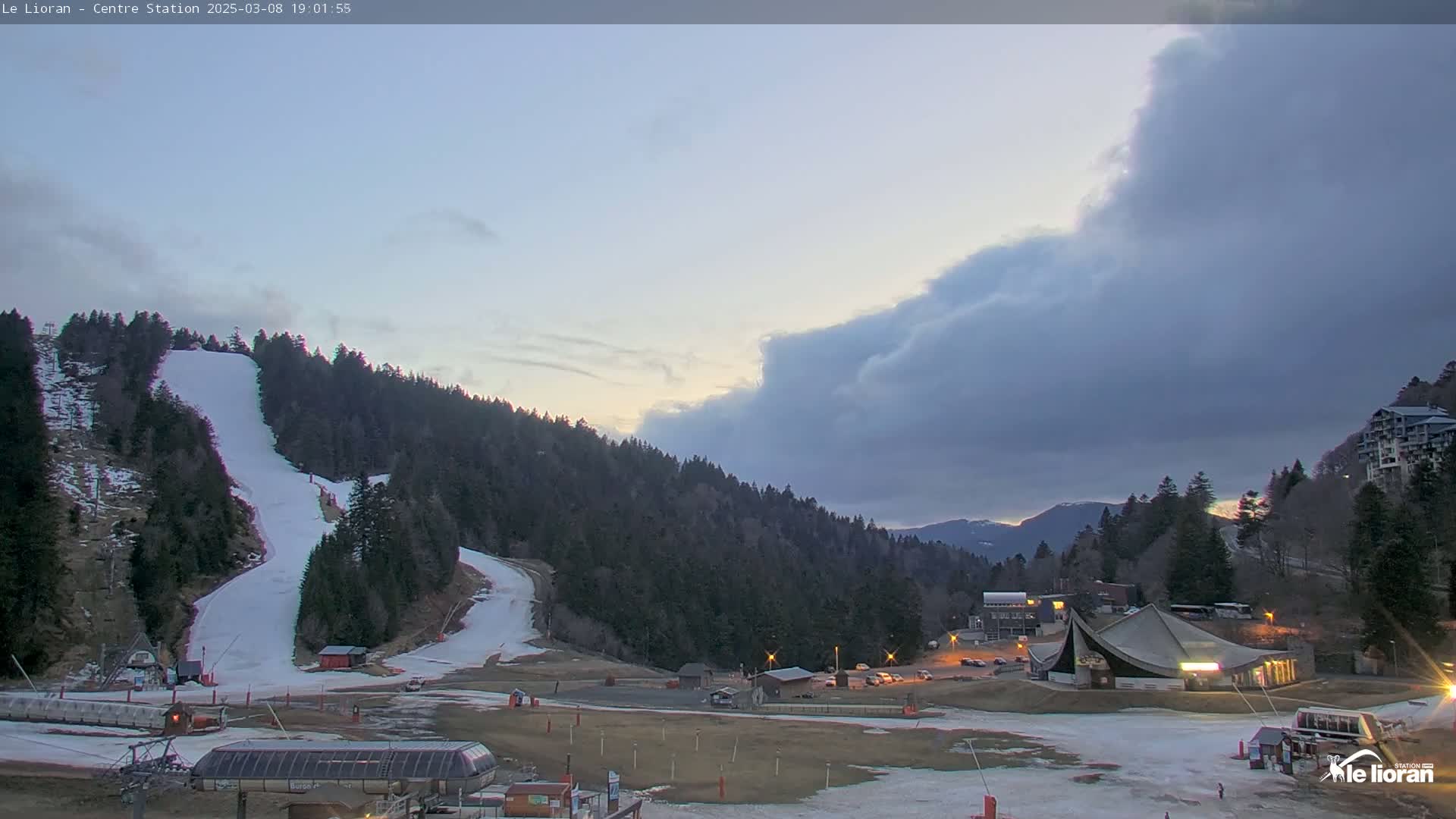 A ski slope with snow and several buildings at the base is visible under a partly cloudy twilight sky.