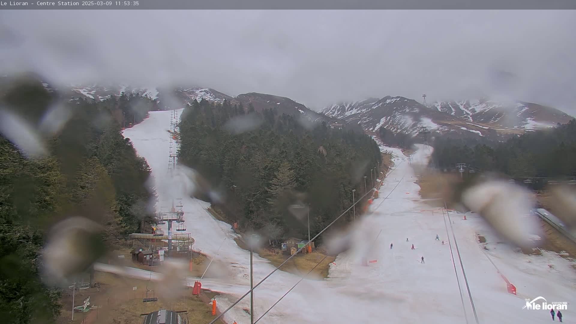 A rain-covered view of a ski slope with several skiers visible on a mostly snow-covered hill, surrounded by dark green trees and mountains under an overcast sky.