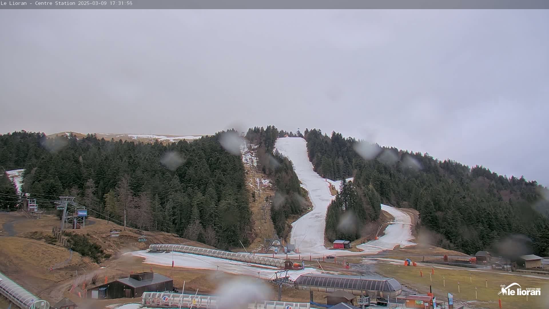 A ski slope with snow and some bare patches is visible through rain-covered glass, surrounded by evergreen trees and ski resort infrastructure.