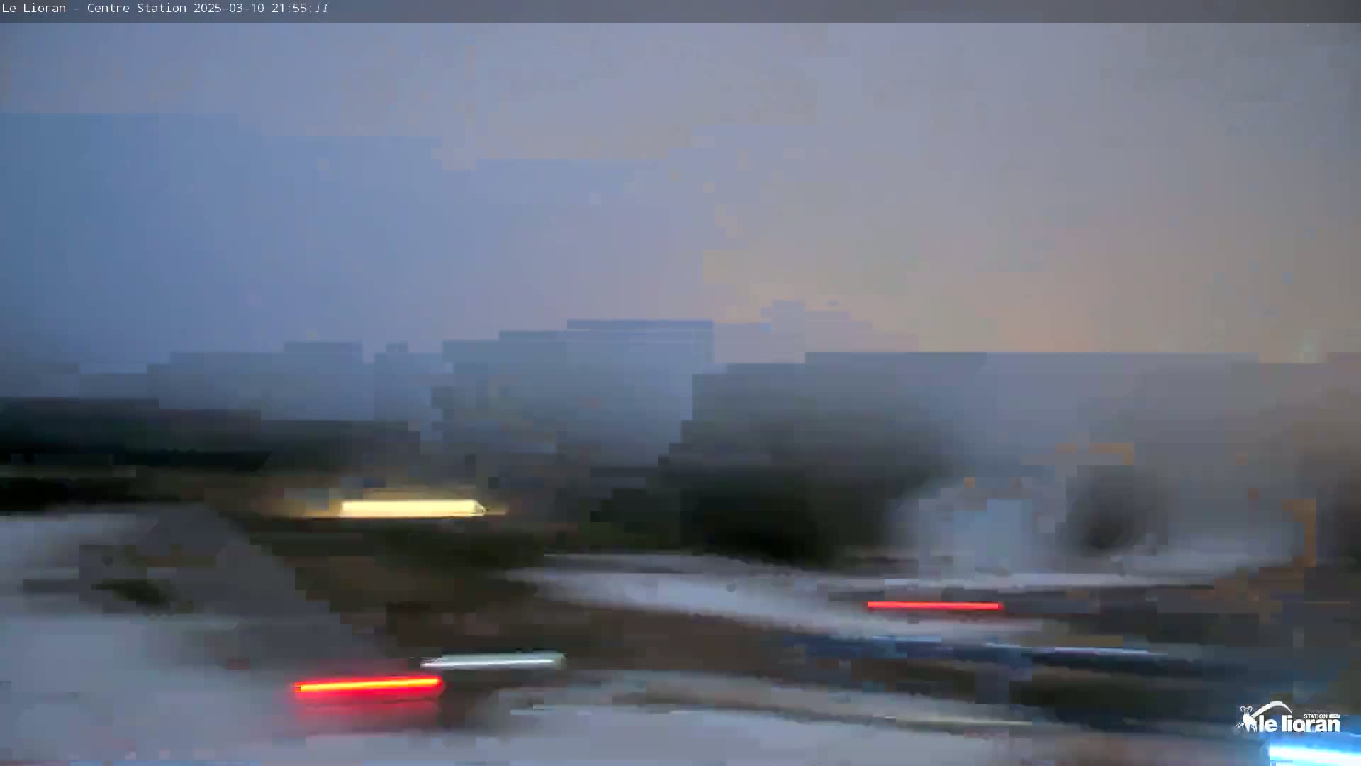 A blurry image shows a hazy, dusk scene of a roadway with indistinct buildings in the background and the tail lights of vehicles visible on the road.