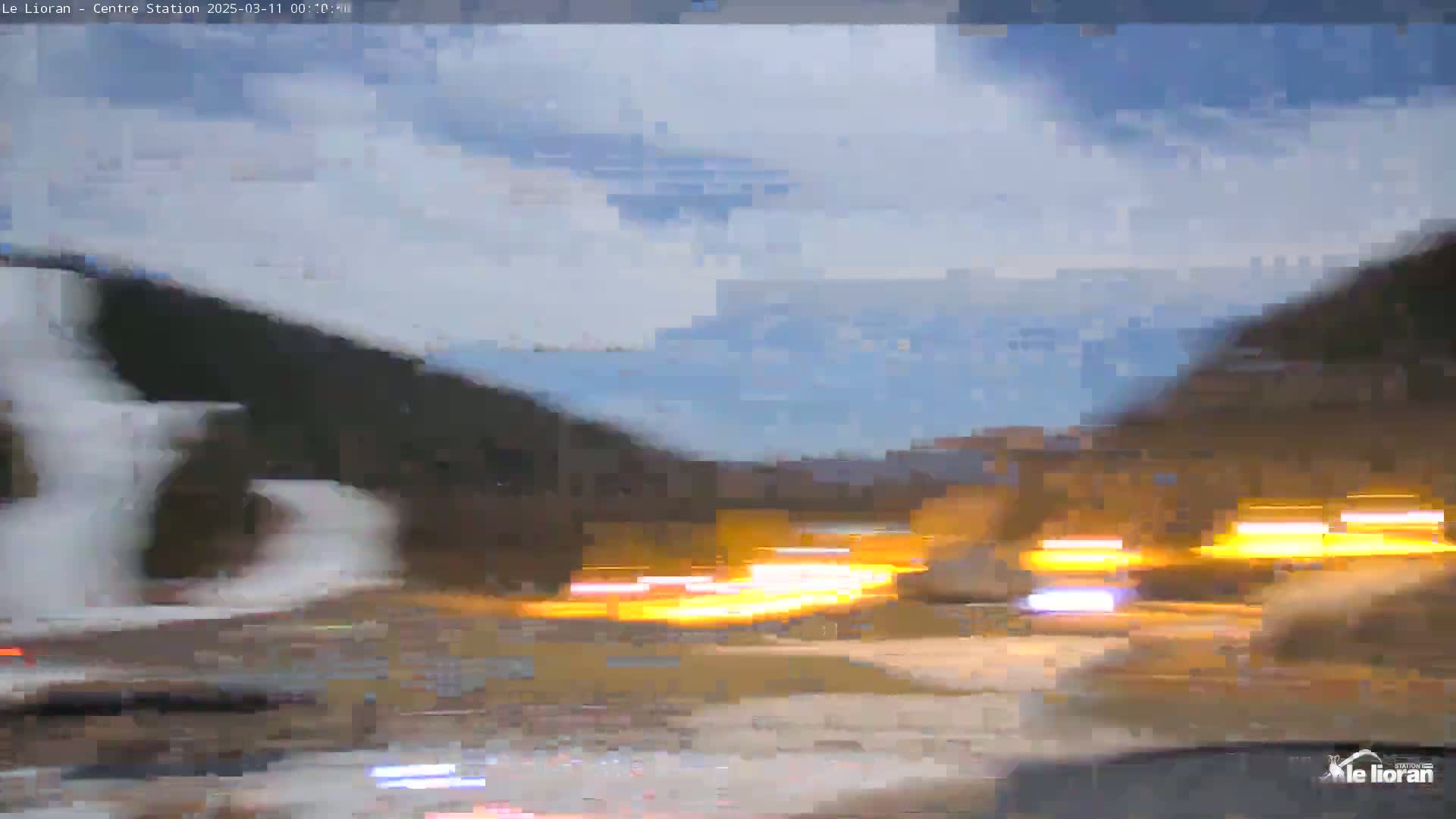 A blurry view of headlights from oncoming traffic on a road at dusk or dawn, with dark hills and a partly cloudy sky in the background.
