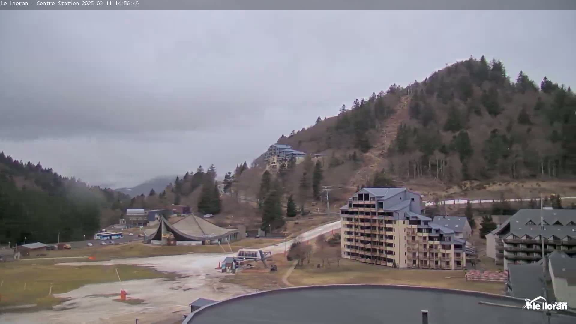 A view of a mountainous region on an overcast day shows several buildings, a ski lift, and a mostly empty ski slope.