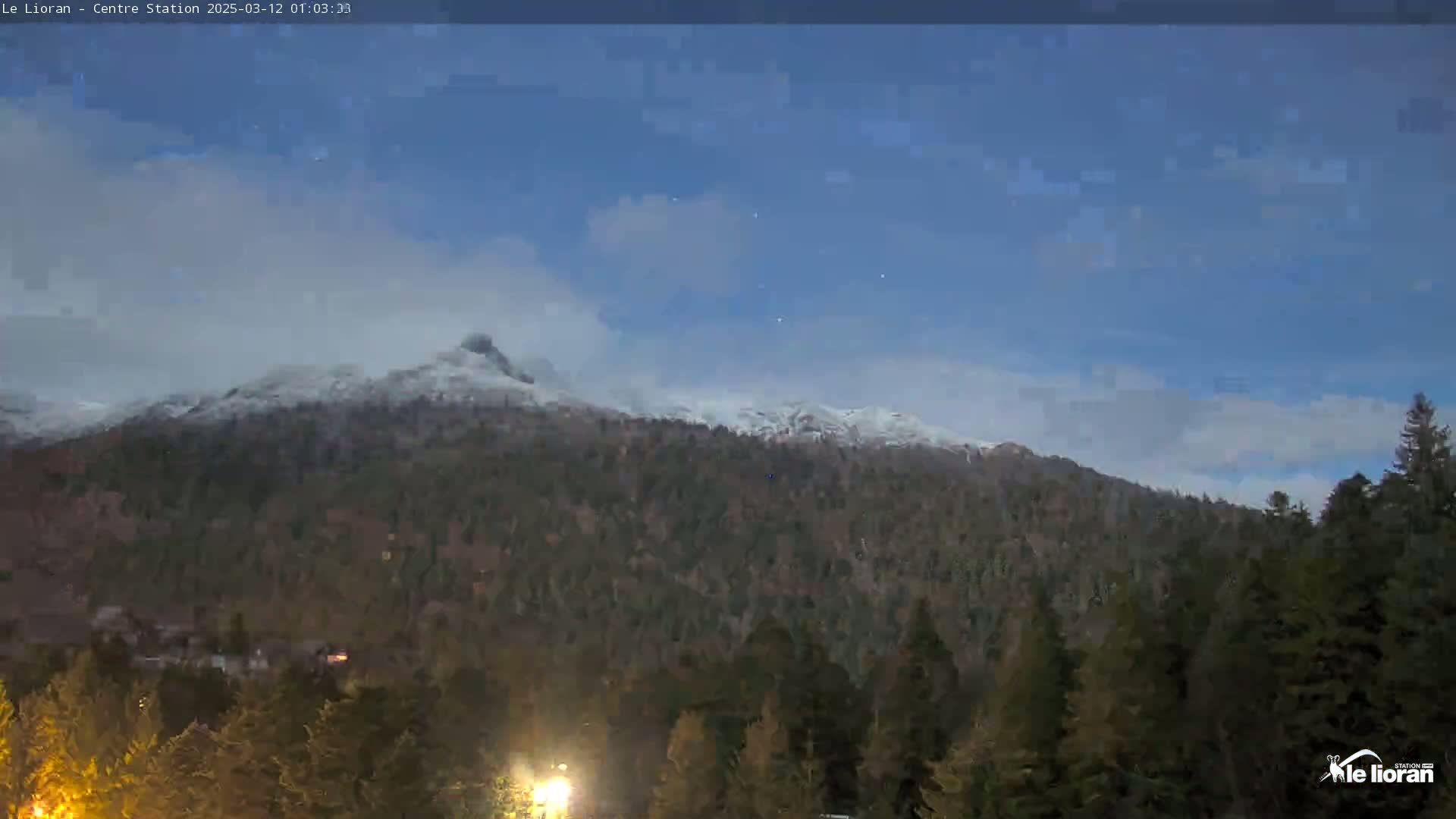 A snow-dusted mountain range is visible under a mostly clear twilight sky above a dark evergreen forest.