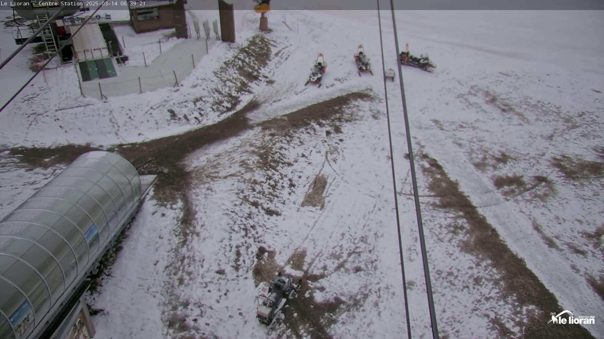 A snow-covered area with several snowmobiles parked near a ski lift station, under an overcast sky.