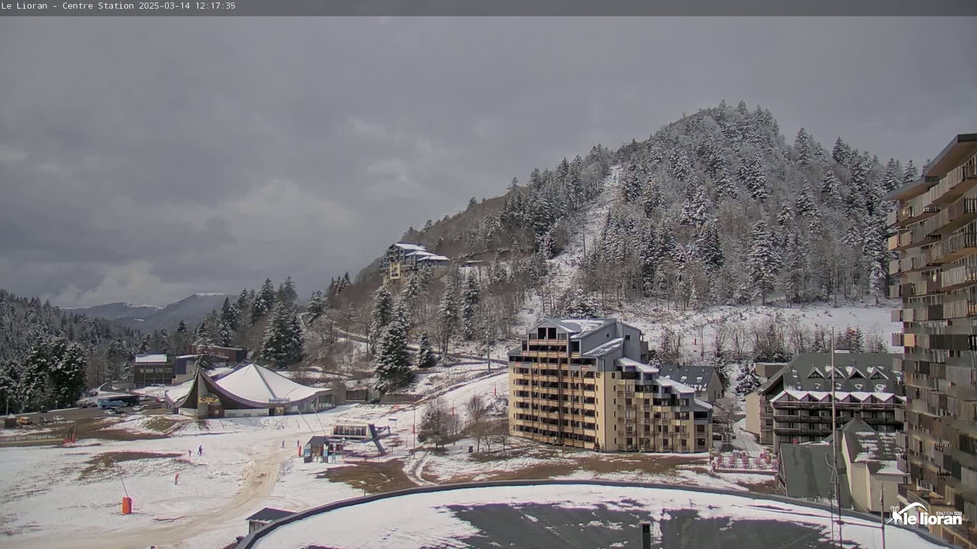 A snow-covered mountainside with several buildings and a ski slope visible under a cloudy sky.