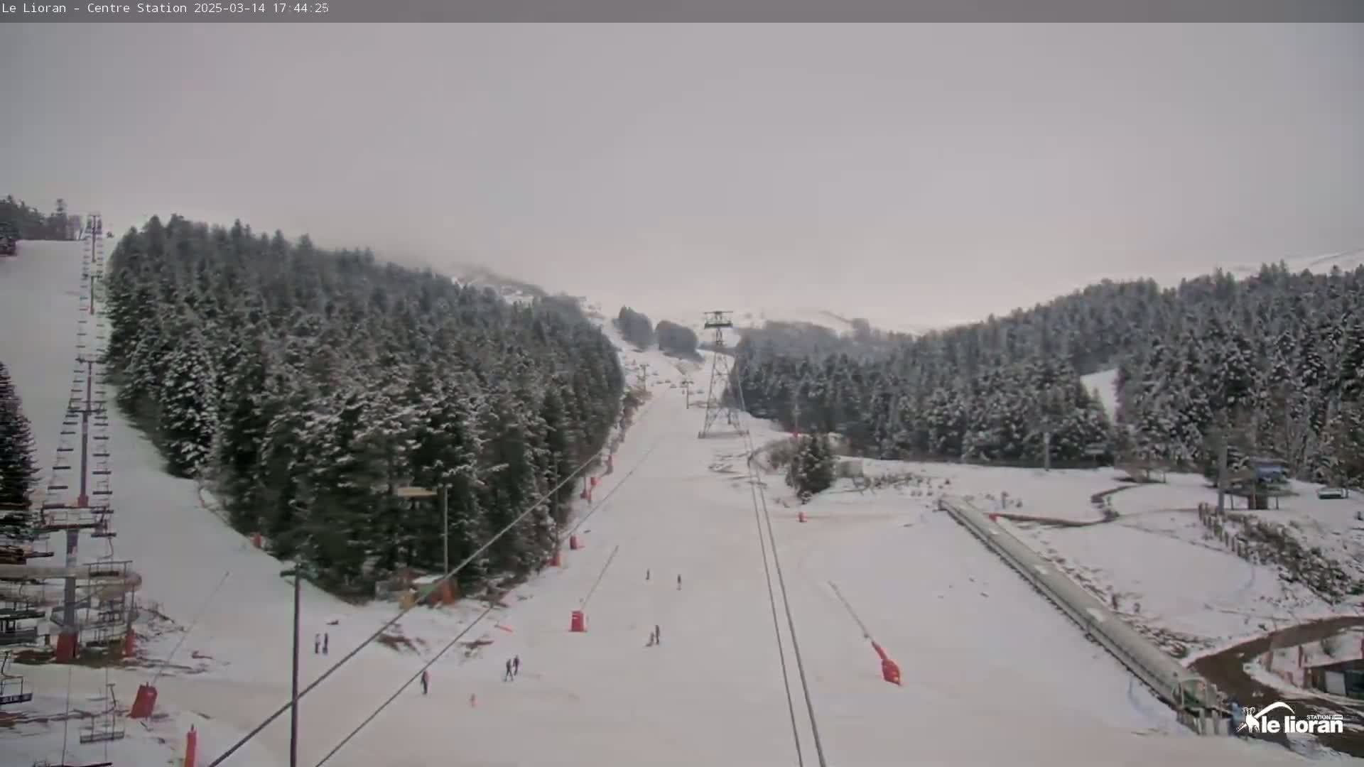 A snow-covered ski slope with a chairlift and several skiers visible under an overcast sky.