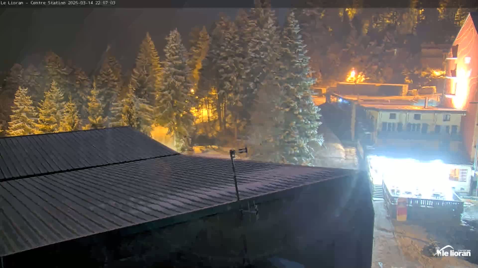 A nighttime view shows snow-covered evergreen trees, buildings with lights, and a dark-colored roof with a small antenna, all under snowy conditions.
