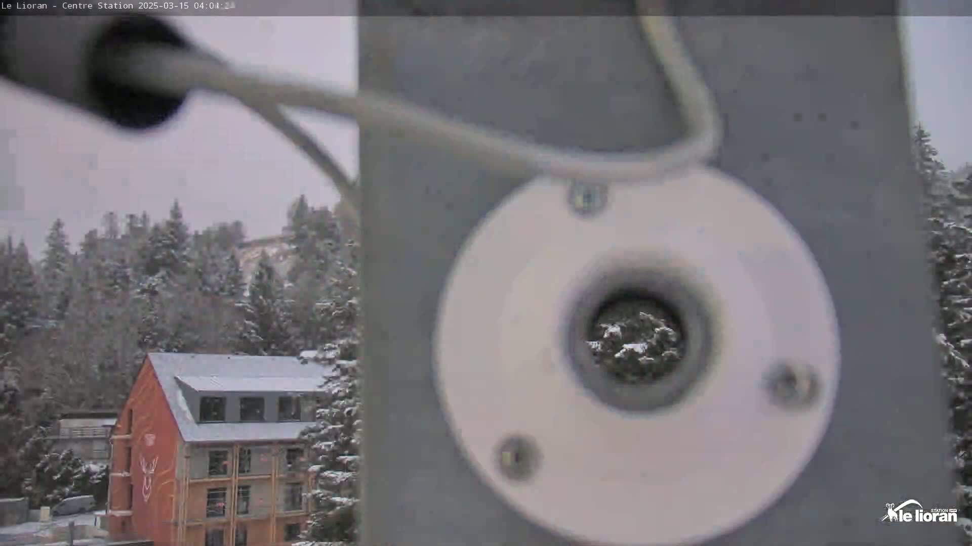 A snow-covered landscape with a multi-story orange building is visible through a partially obscured view from a gray pole with a circular attachment.