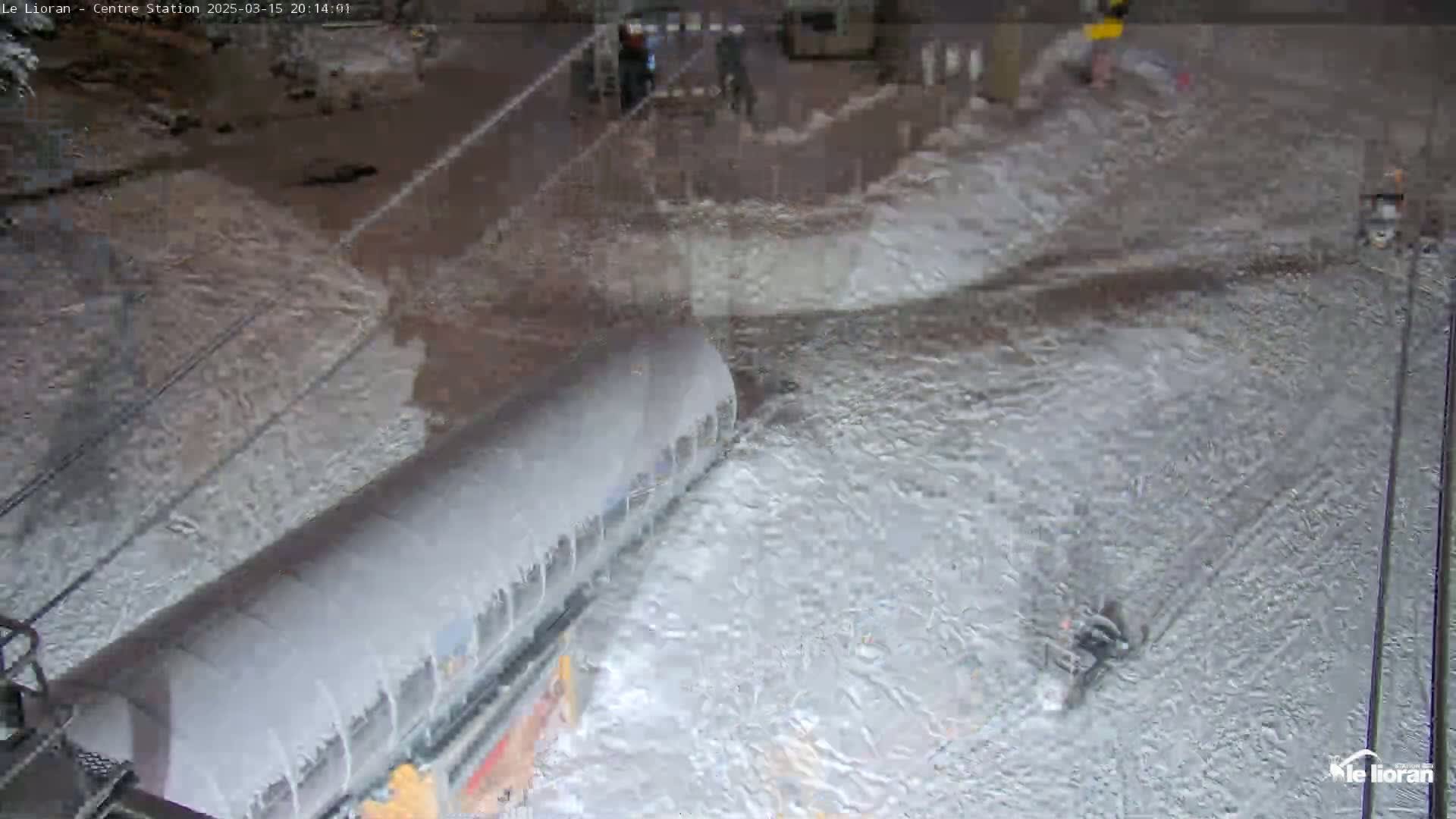 A snow-covered cable car sits on a snowy ground with several people in the distance under nighttime conditions.