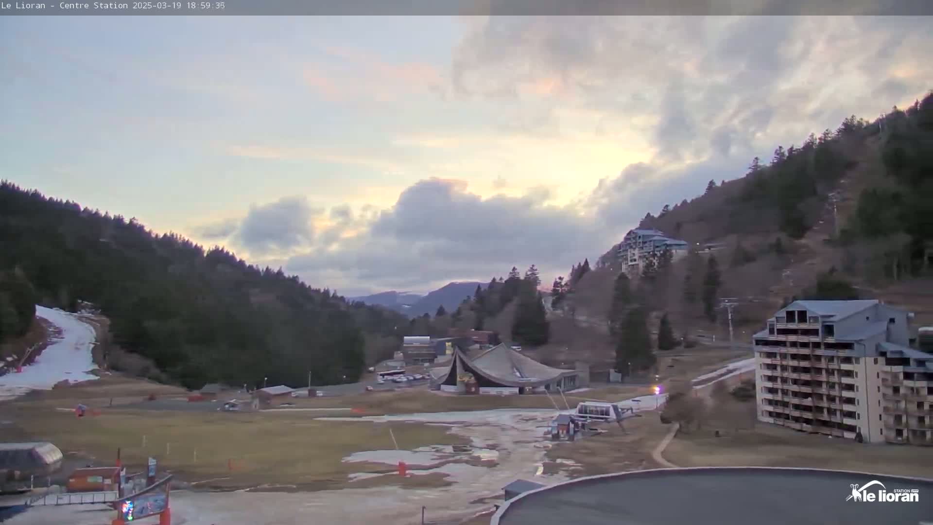 A mountain valley scene at dusk shows partly cloudy skies, snow patches on the ground, and several buildings nestled amongst the trees.