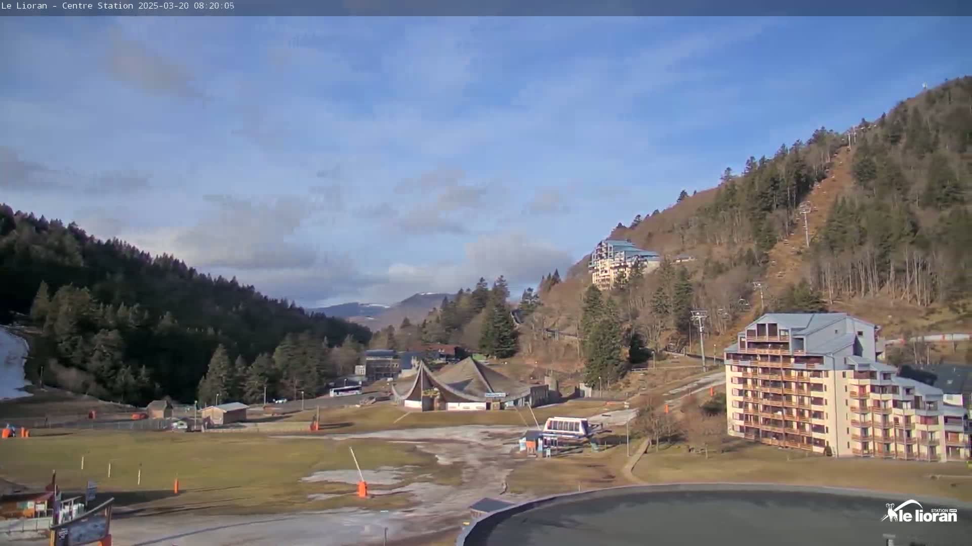 A mostly sunny day reveals a ski resort nestled in a valley between snow-dusted mountains, with a large building and chairlifts visible.
