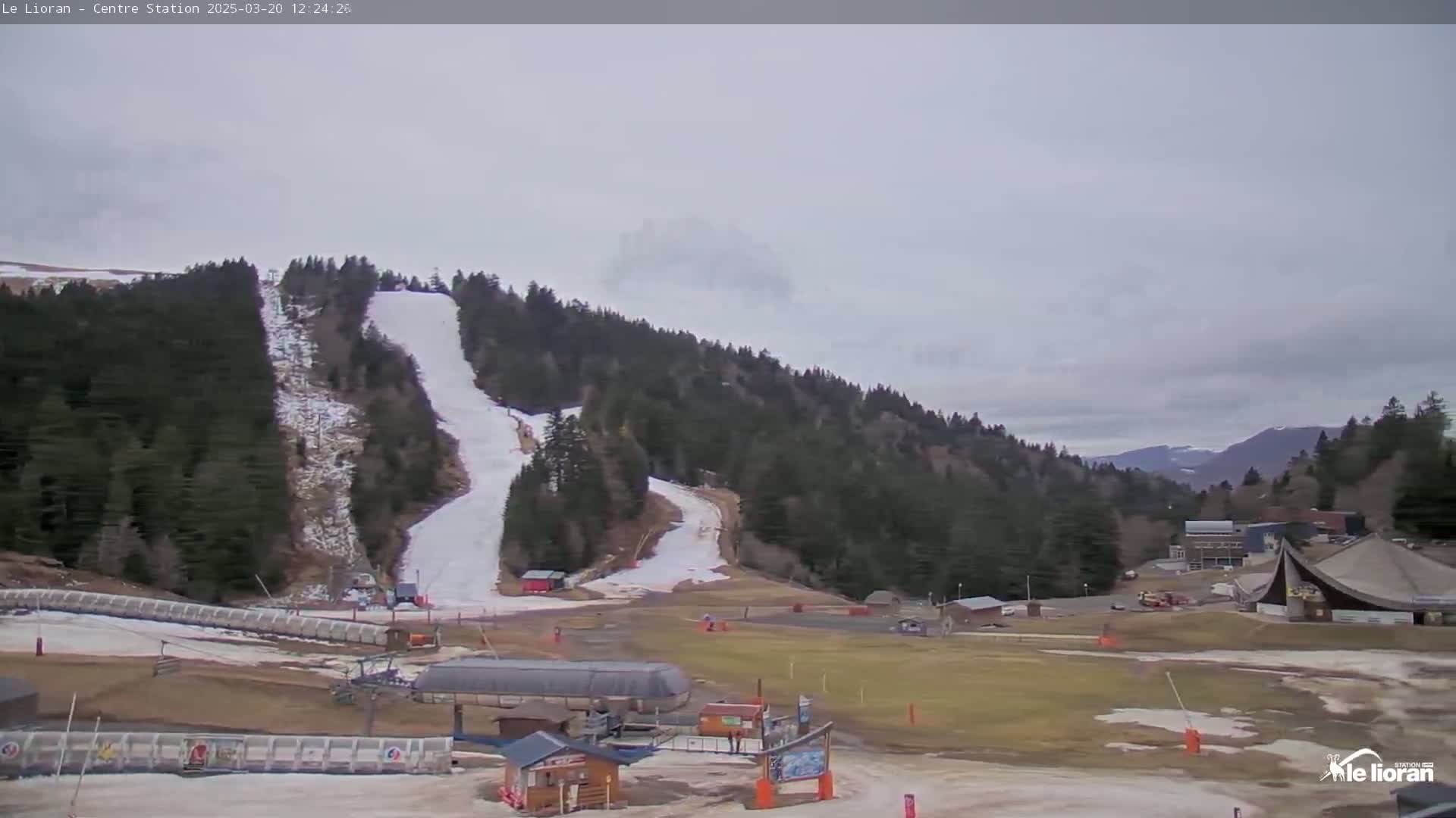 A ski slope with patchy snow on the hill and brown grass in the lower areas, under an overcast sky.