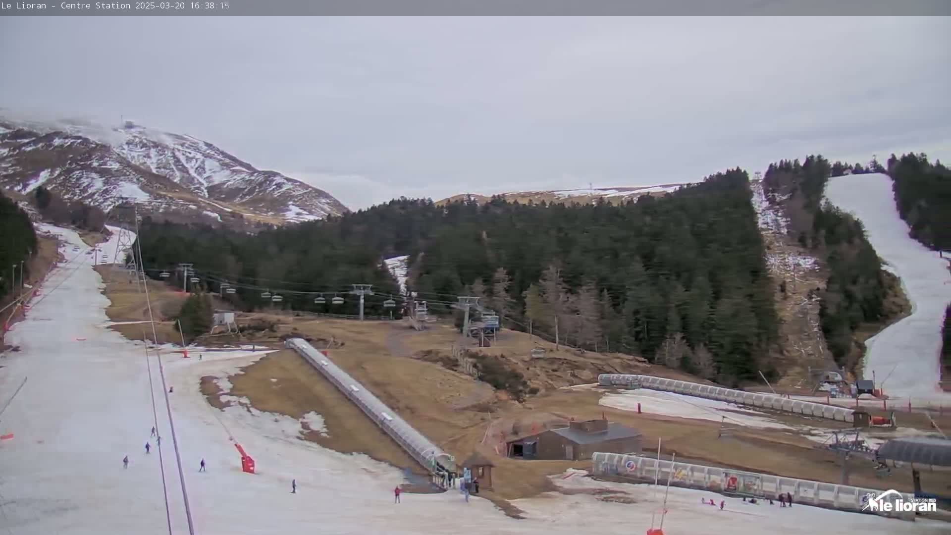 A ski resort on a partly cloudy day shows several ski slopes with skiers, chairlifts, and snow-making equipment.