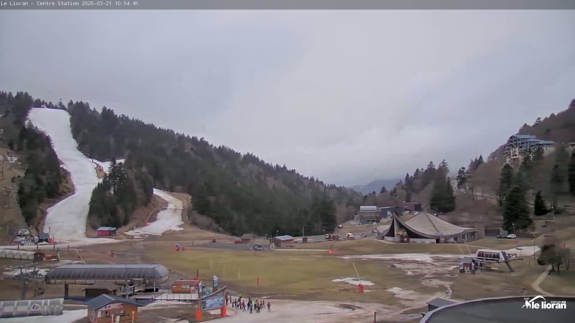 A ski slope with a few people at the bottom, surrounded by snow-covered hills and evergreen trees under an overcast sky.