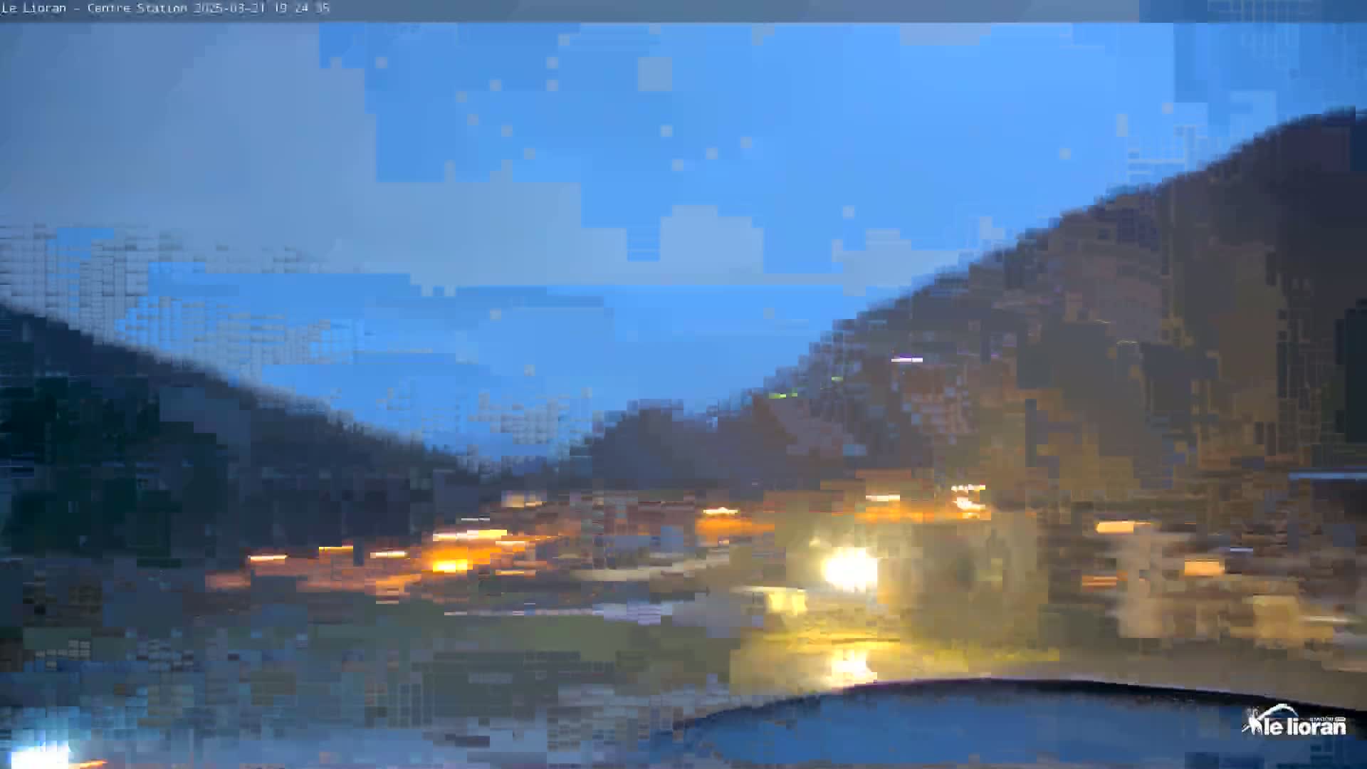 A blurry nighttime view shows a valley with dark hills under a twilight blue sky, illuminated by headlights and scattered lights below.