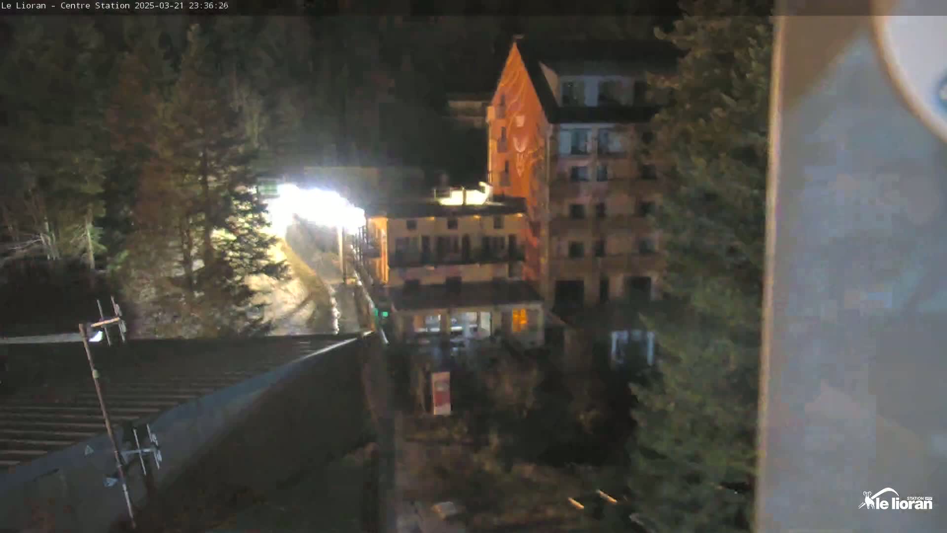 A nighttime, high-angle view of a wet, dimly lit street scene featuring several buildings, trees, and a roadway.