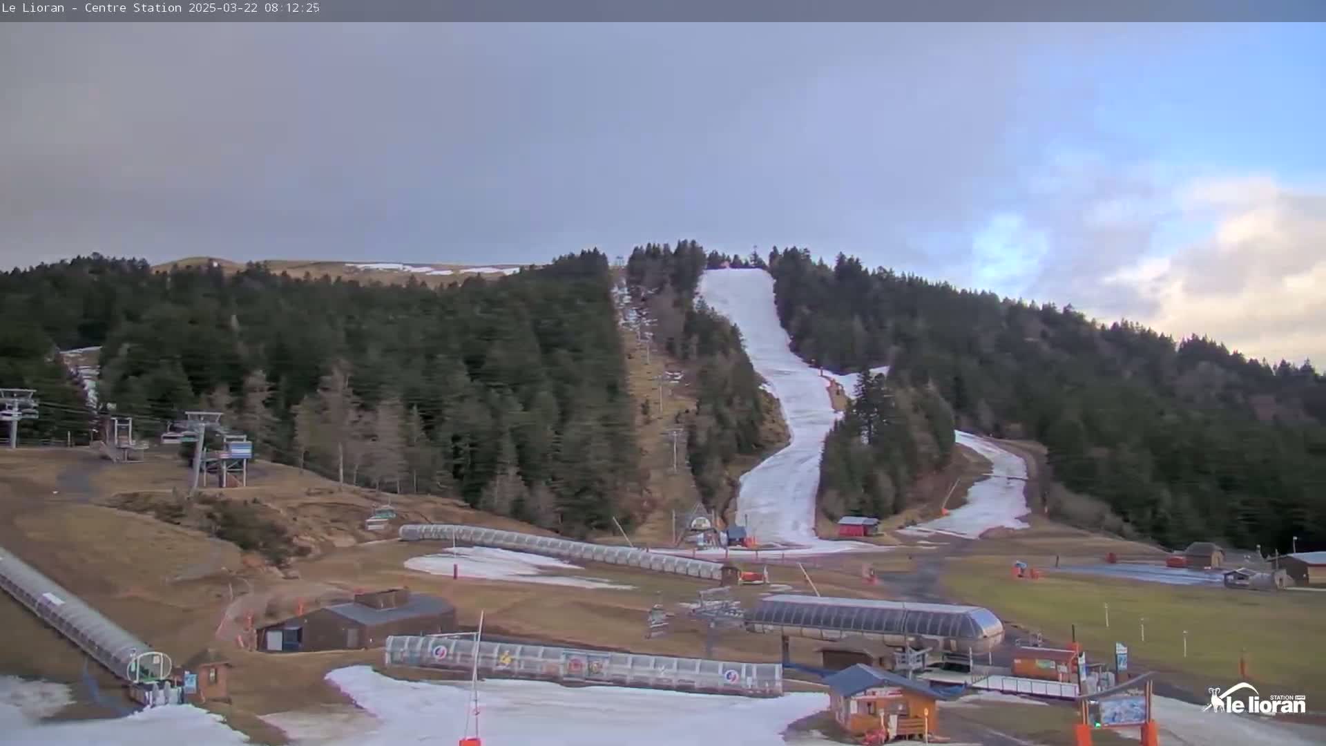 A ski slope with patches of snow and some grassy areas is visible, surrounded by a coniferous forest under a partly cloudy sky.