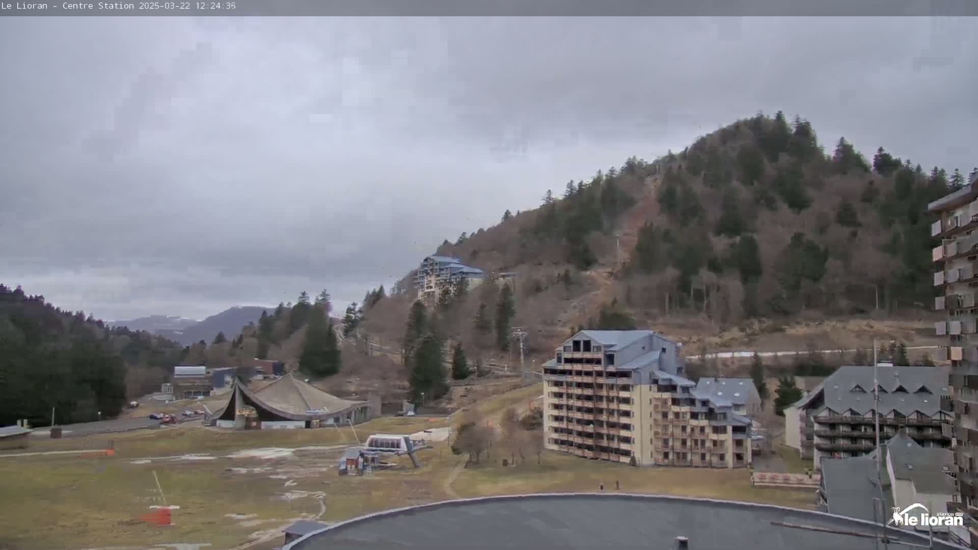 A cloudy day reveals a mountainside dotted with buildings and ski lifts, overlooking a flat expanse of land with sparse vegetation.
