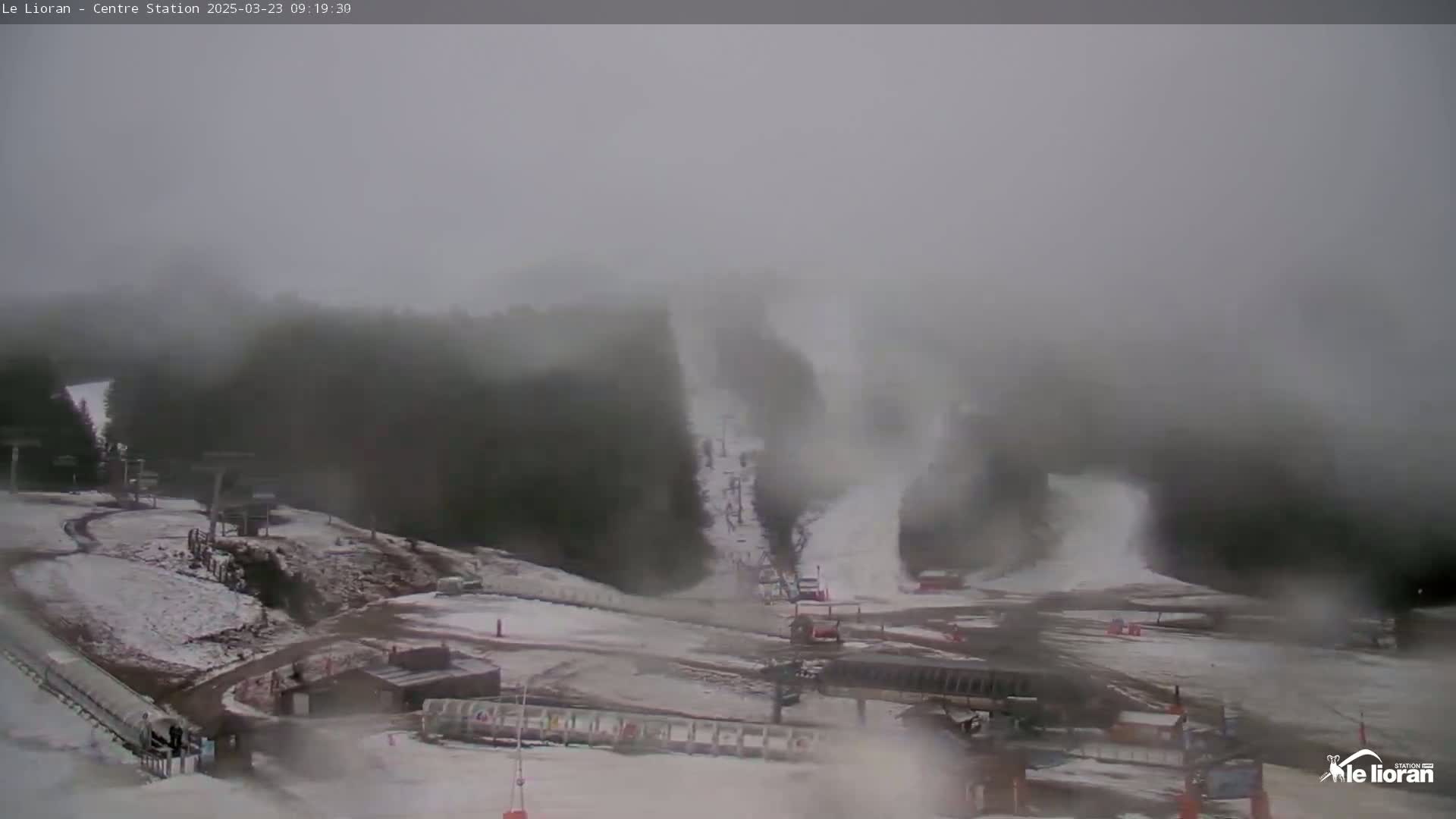 A snow-covered ski resort is shrouded in thick fog, with snow blowing across a ski slope and chairlifts visible in the distance.