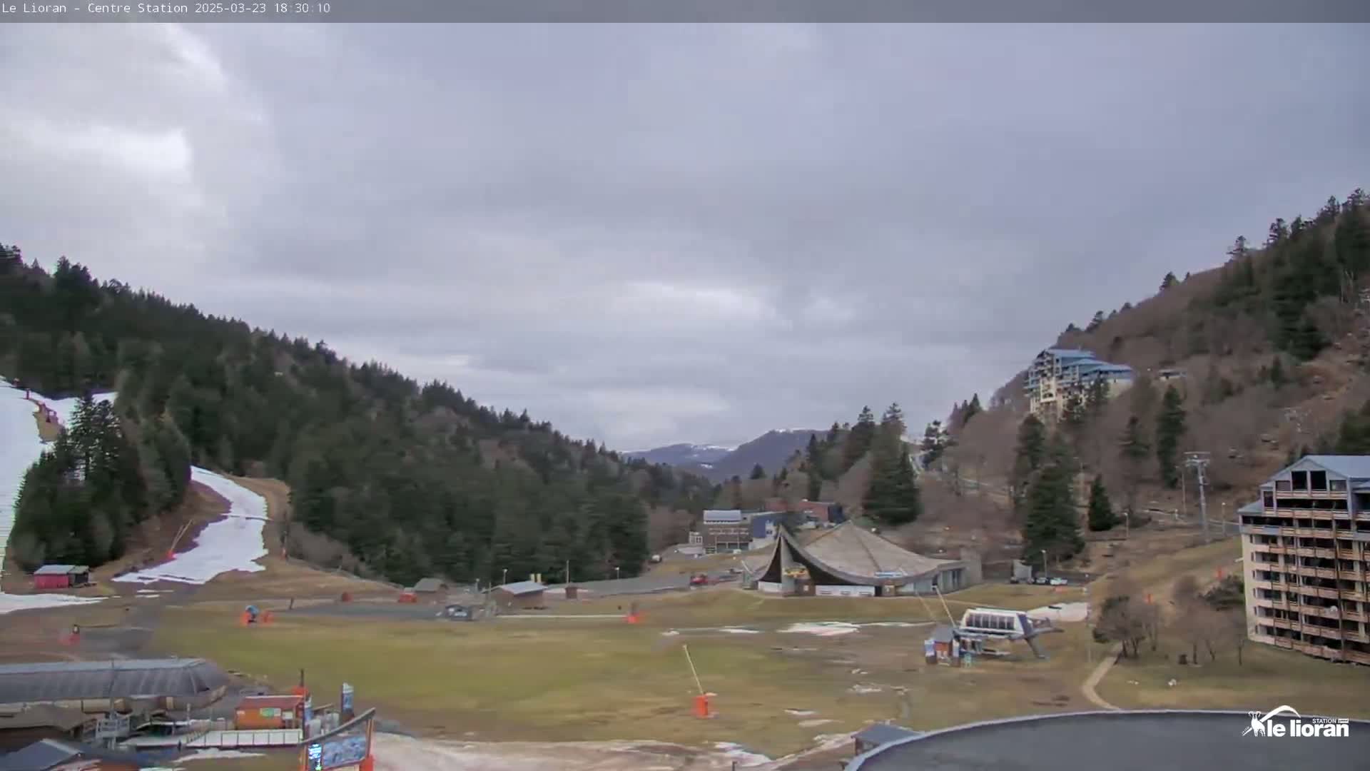 A mostly cloudy day reveals a ski resort nestled in a valley between snow-dusted mountains, with some patches of snow visible on the slopes and in the valley below.