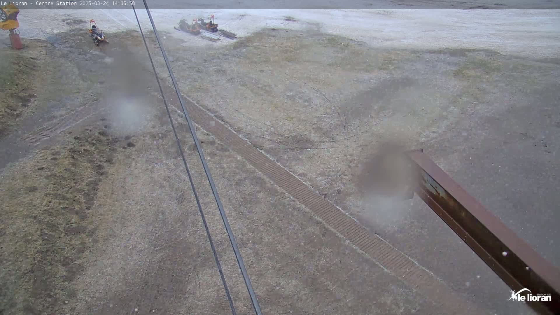 A snow-dusted, sparsely vegetated ground area is seen from an elevated perspective, showing a dirt path and some machinery in the distance under hazy conditions.