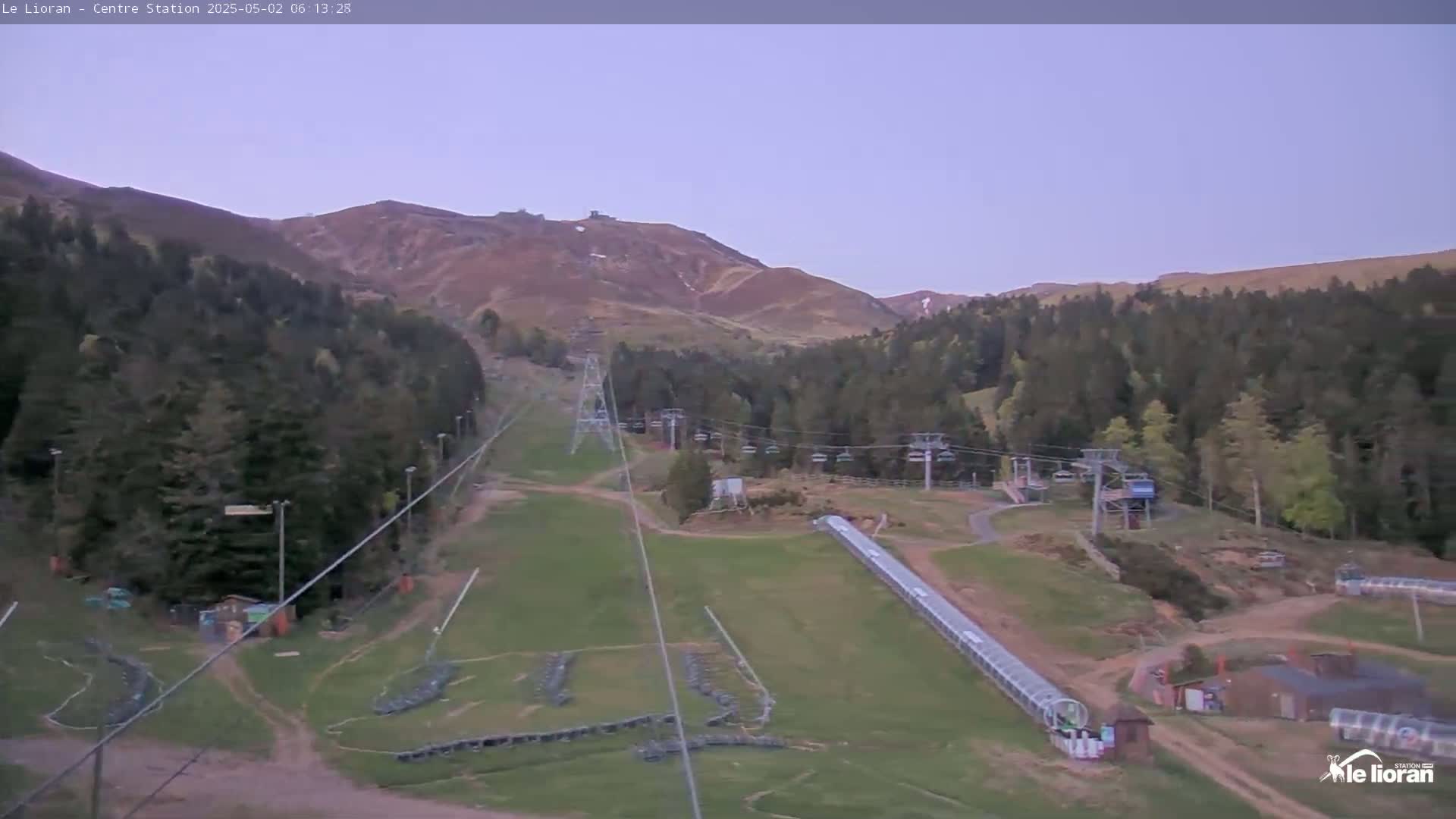 A ski resort in the mountains on a partly cloudy day shows chairlifts, grassy slopes, and a covered ski lift.