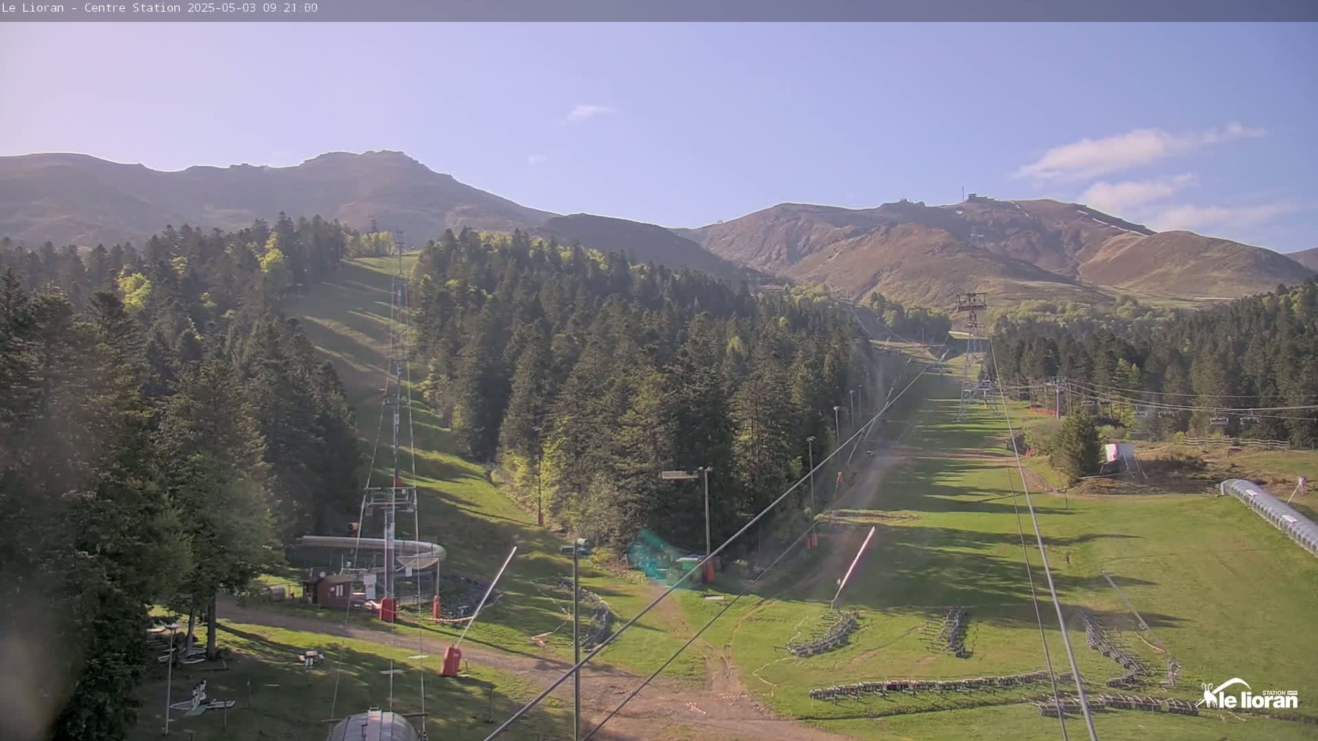 A sunny view of a ski resort in the mountains, showing grassy slopes, ski lifts, and evergreen trees.
