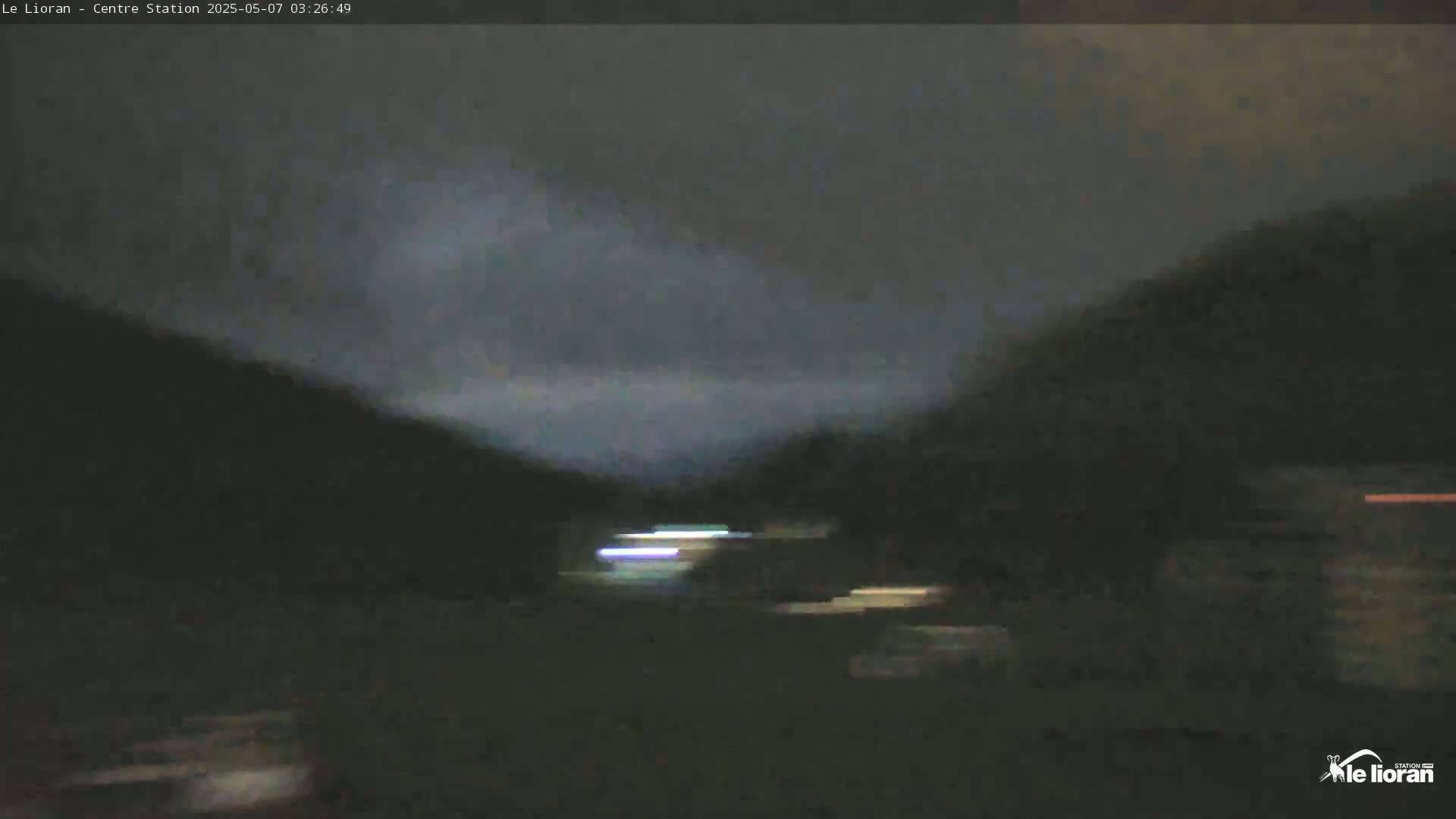 A dark, overcast night scene shows a blurry view of a road with faint lights in the distance, surrounded by dark hills.