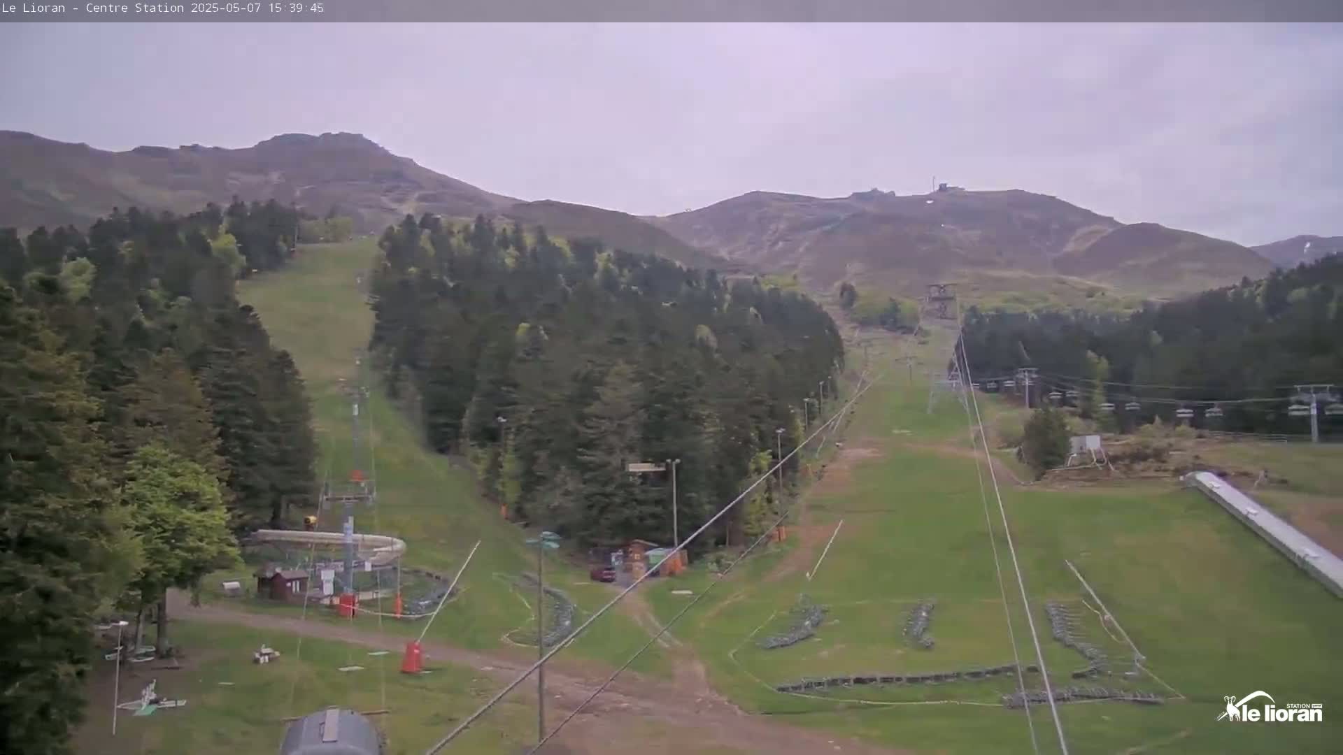 A ski slope with chairlifts is visible, surrounded by a mountainous area covered in green forests under an overcast sky.