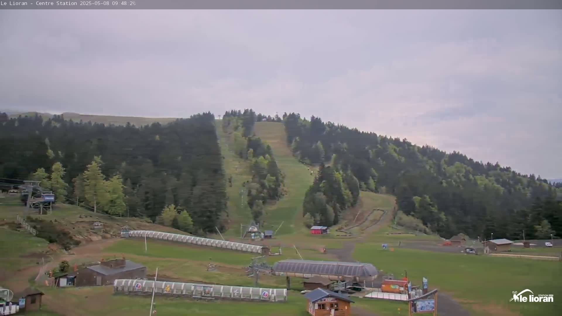 A ski resort in a mostly cloudy day shows several ski lifts, grassy areas, and a hill covered with evergreen trees.