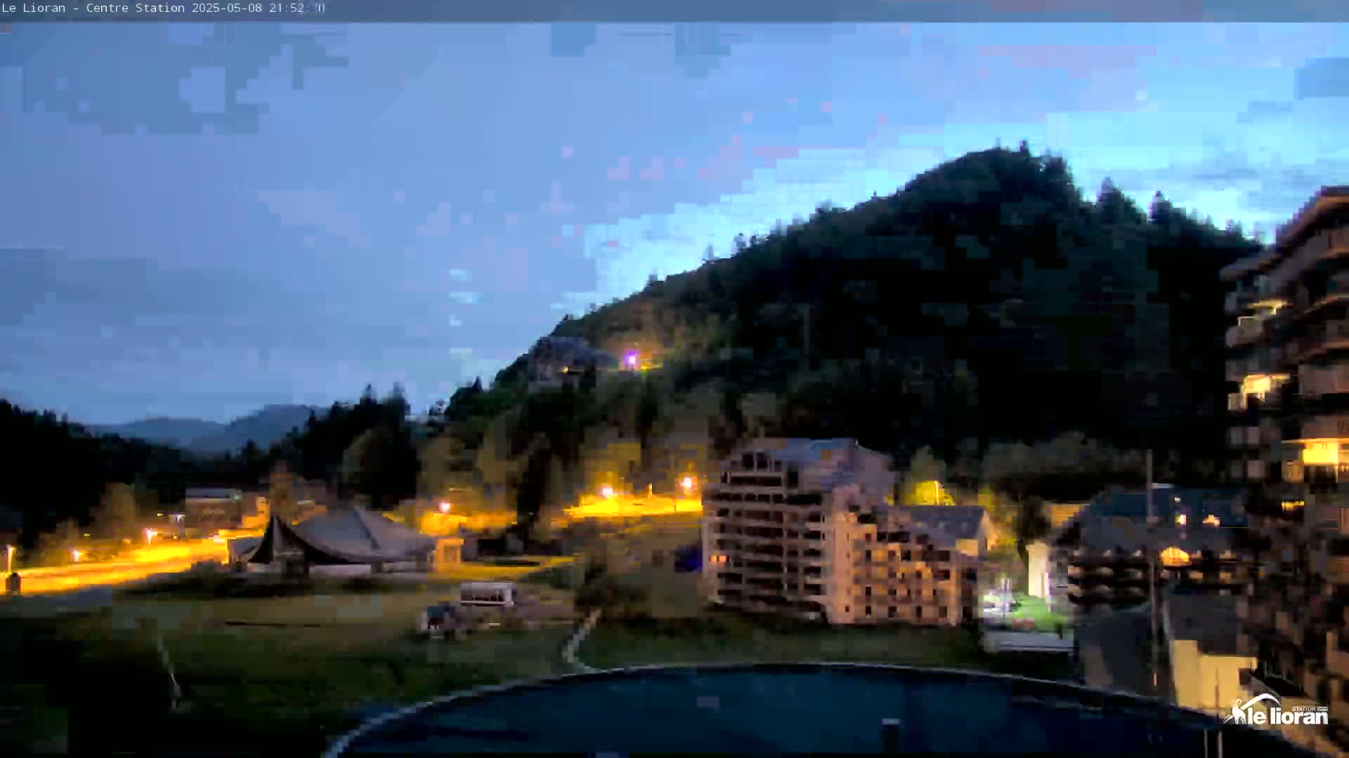 A nighttime view of a mountainside town with illuminated buildings and roads under a twilight sky.