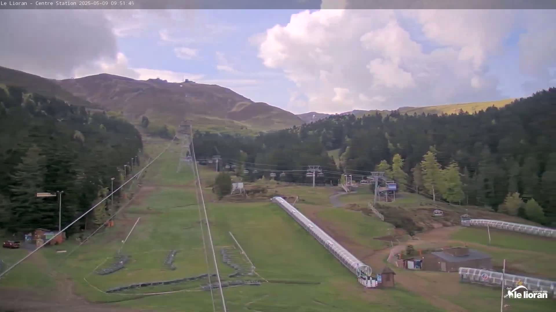 A partly cloudy sky overlooks a ski resort with chairlifts, a covered ski lift, and green grassy slopes surrounded by dark green coniferous forests and brown hills.