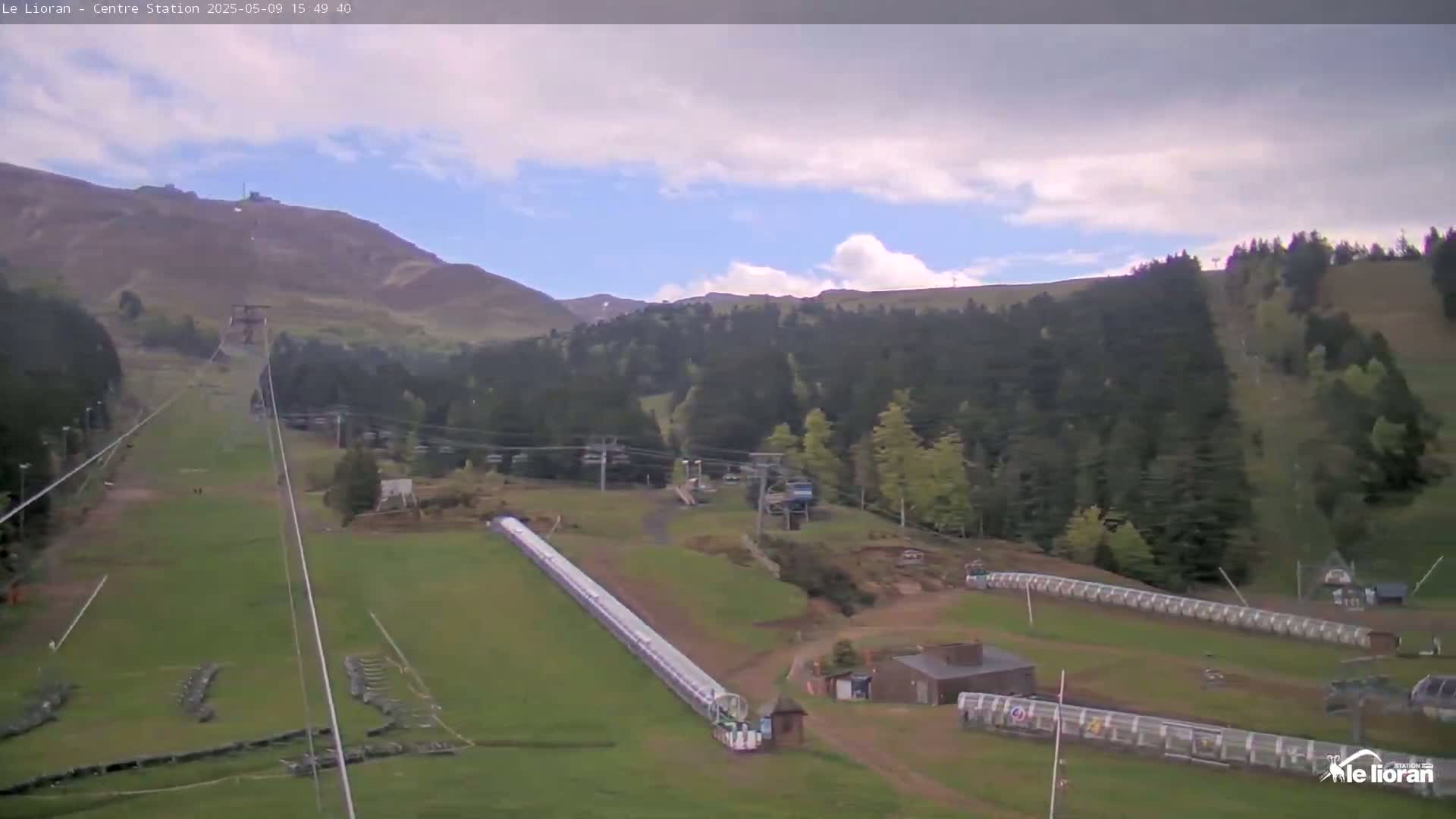 A long, grassy ski slope with a chairlift and several buildings is visible beneath a partly cloudy sky.