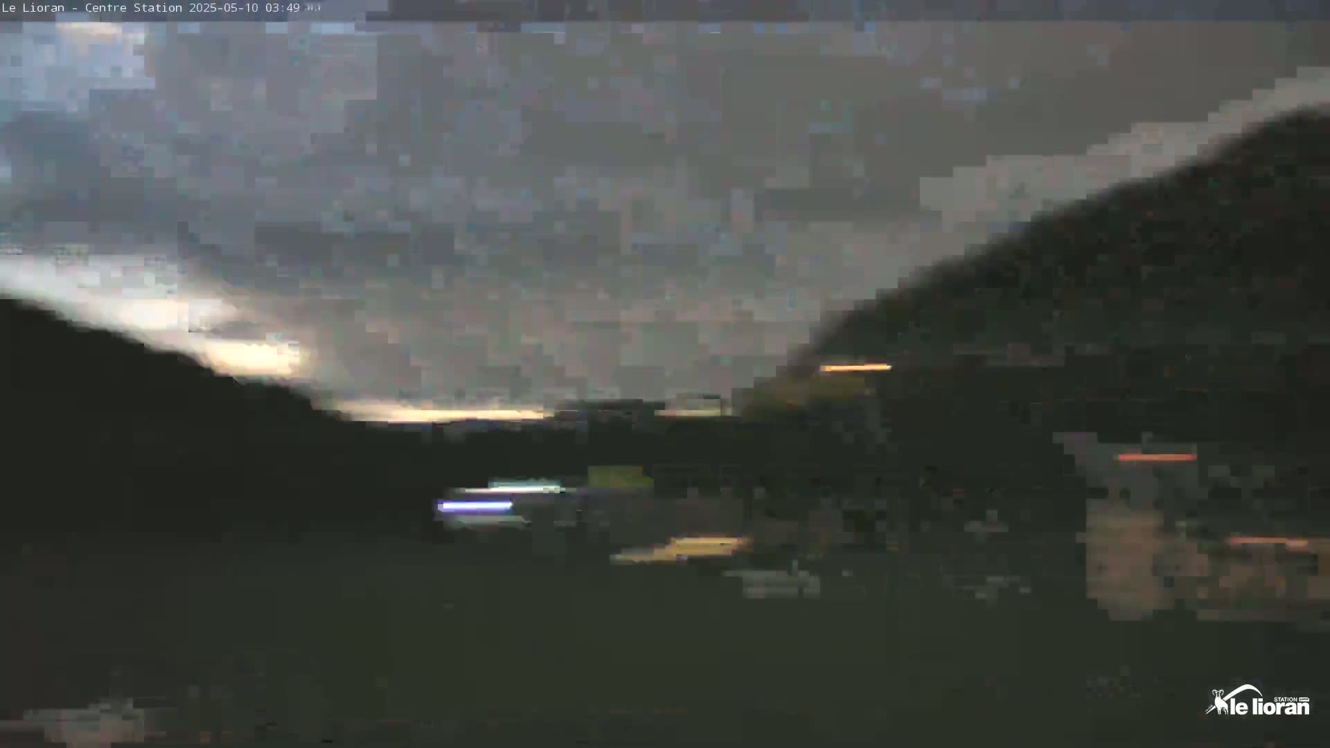 A dark, overcast sky illuminates a mountainous landscape at twilight, with blurry lights visible in the distance.