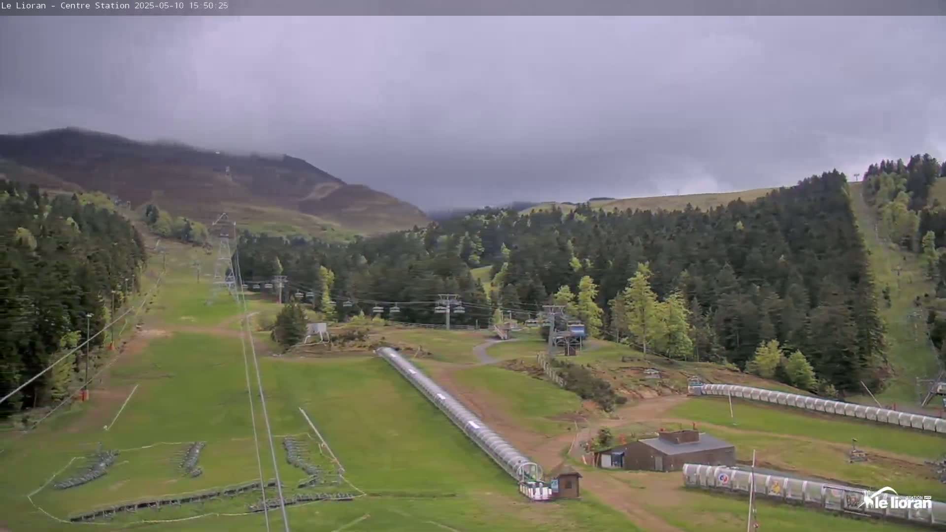 A ski resort in a mountainous area under an overcast sky shows grassy slopes, chairlifts, and covered conveyor belts.