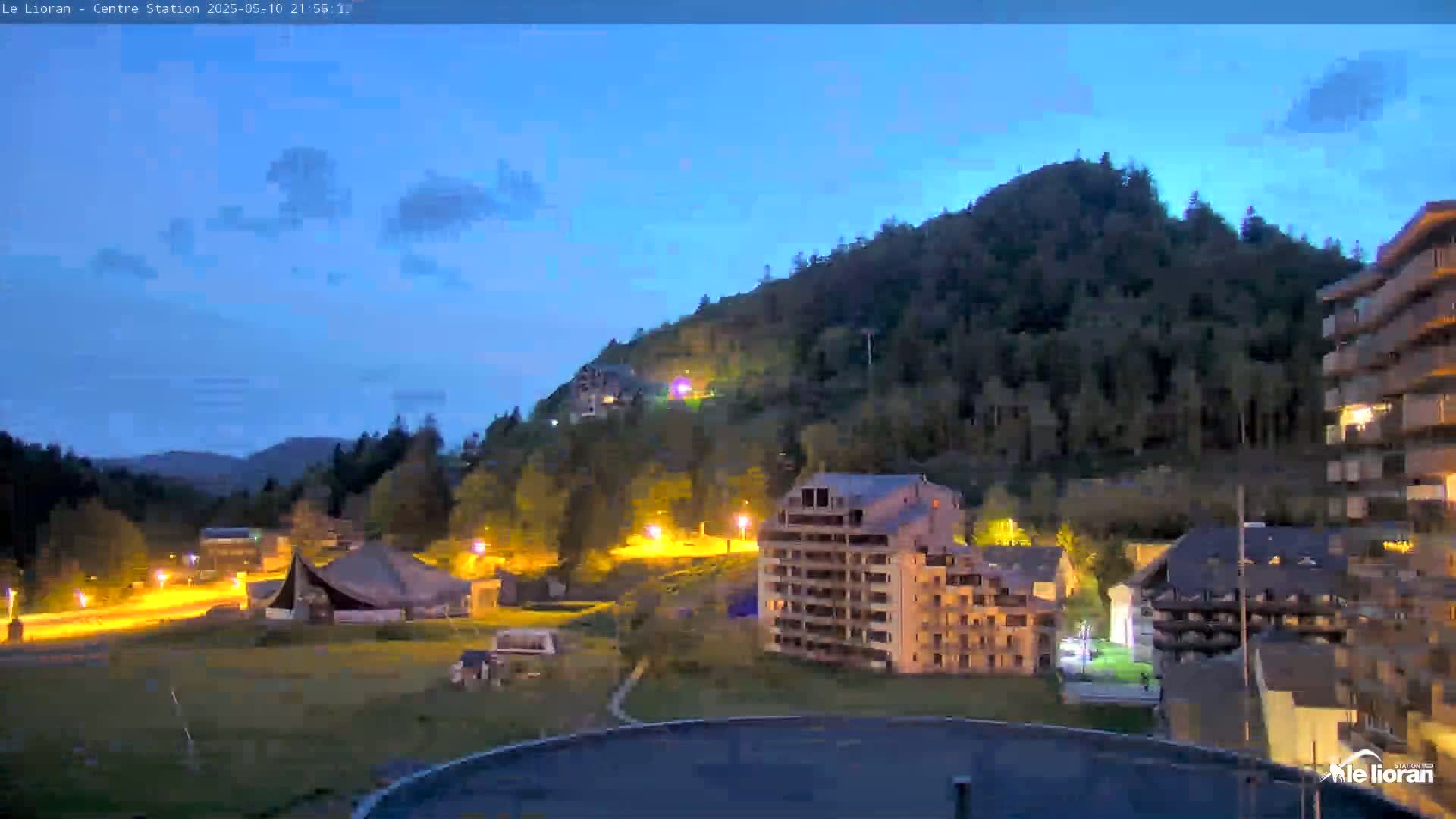 A twilight view of a mountain town illuminated at night, with a clear sky and a few clouds.