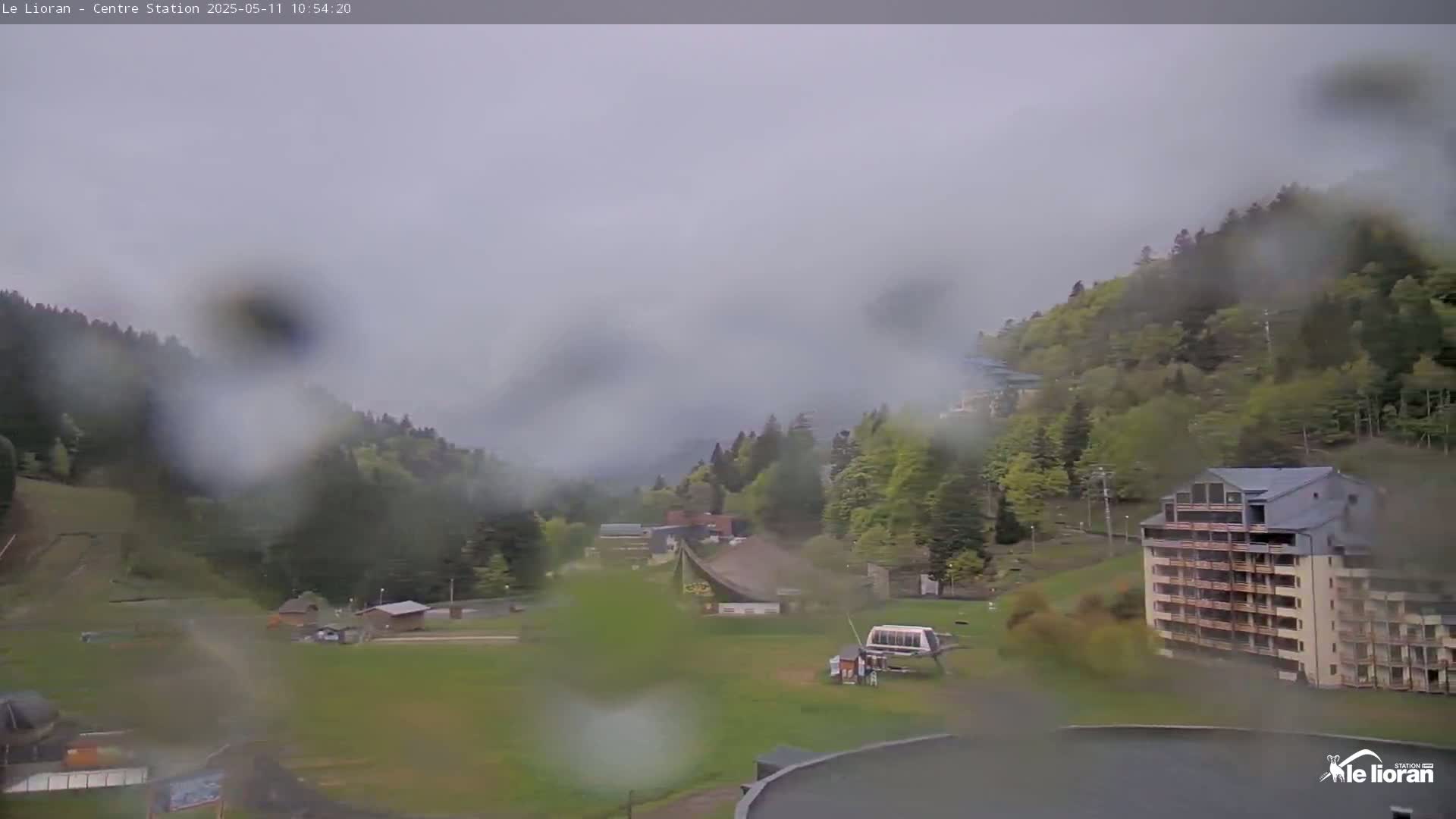 A rain-streaked view shows a grassy valley with buildings, a chairlift, and forested hills under a cloudy sky.