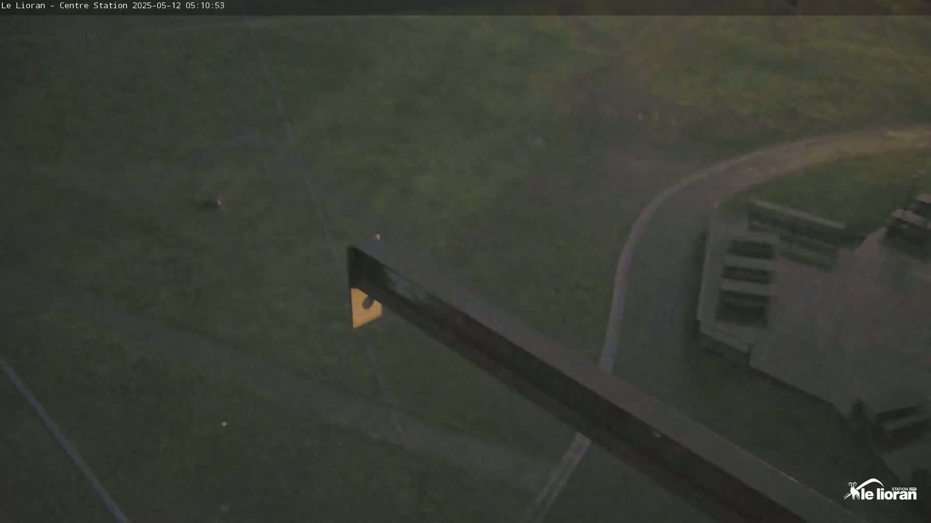 Here is a description of the image: A high-angle, low-light view shows a paved road curving through a grassy area next to a building, all under overcast skies.