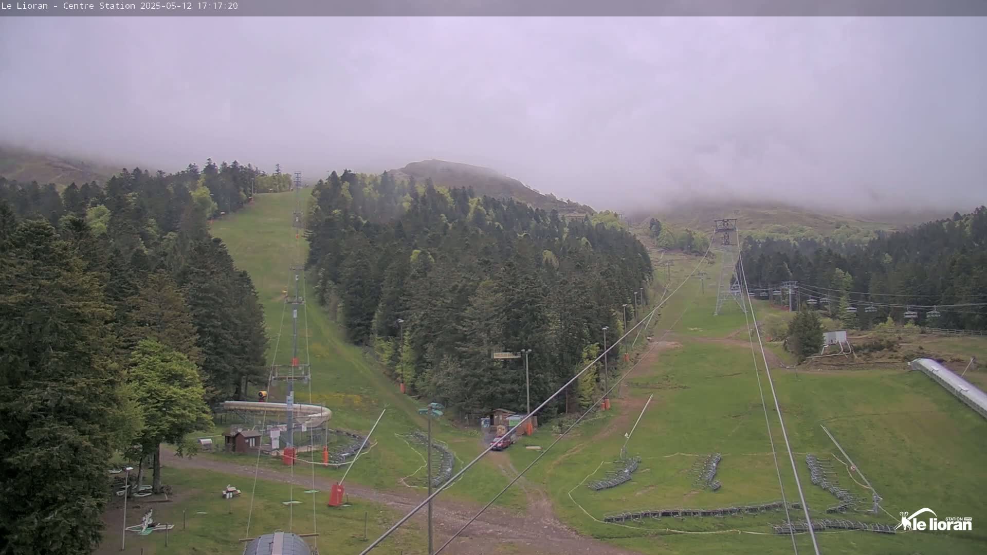 A foggy view of a ski resort with green grassy slopes, chairlifts, and evergreen trees covering the surrounding hills.