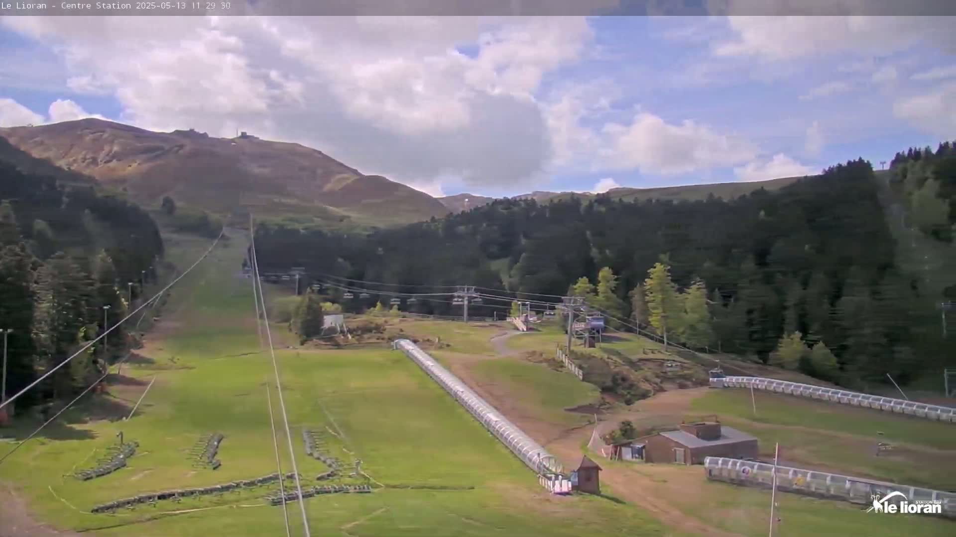 A mostly sunny day reveals a mountain ski resort with chairlifts, grassy slopes, and a covered conveyor lift.