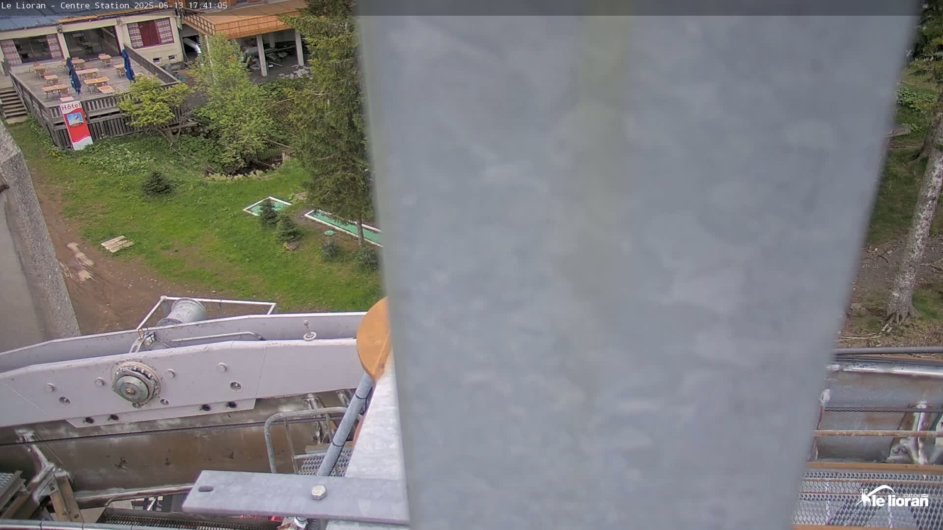 A partly obstructed view of a chairlift mechanism overlooks a grassy area with some small trees and a miniature golf course on a partly cloudy day.