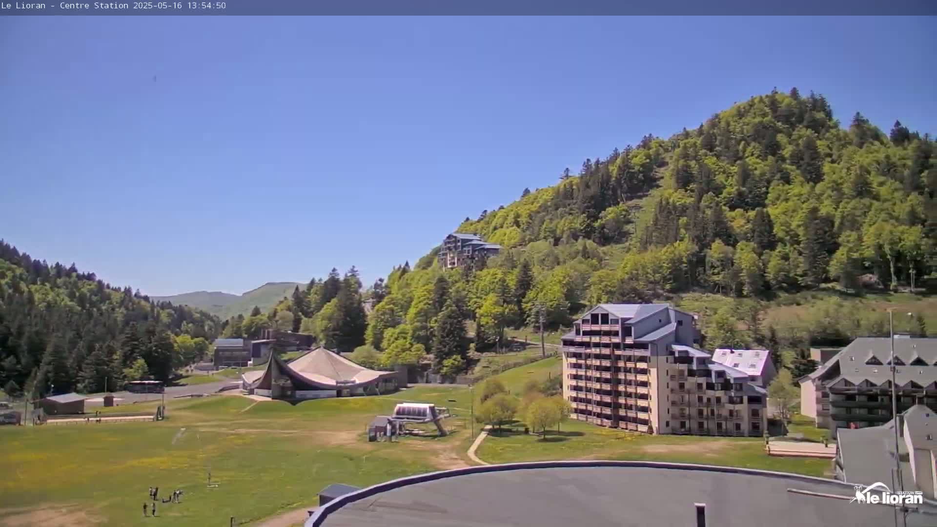 A sunny day reveals a mountainside covered in lush green trees, with several buildings and a grassy area in the foreground.
