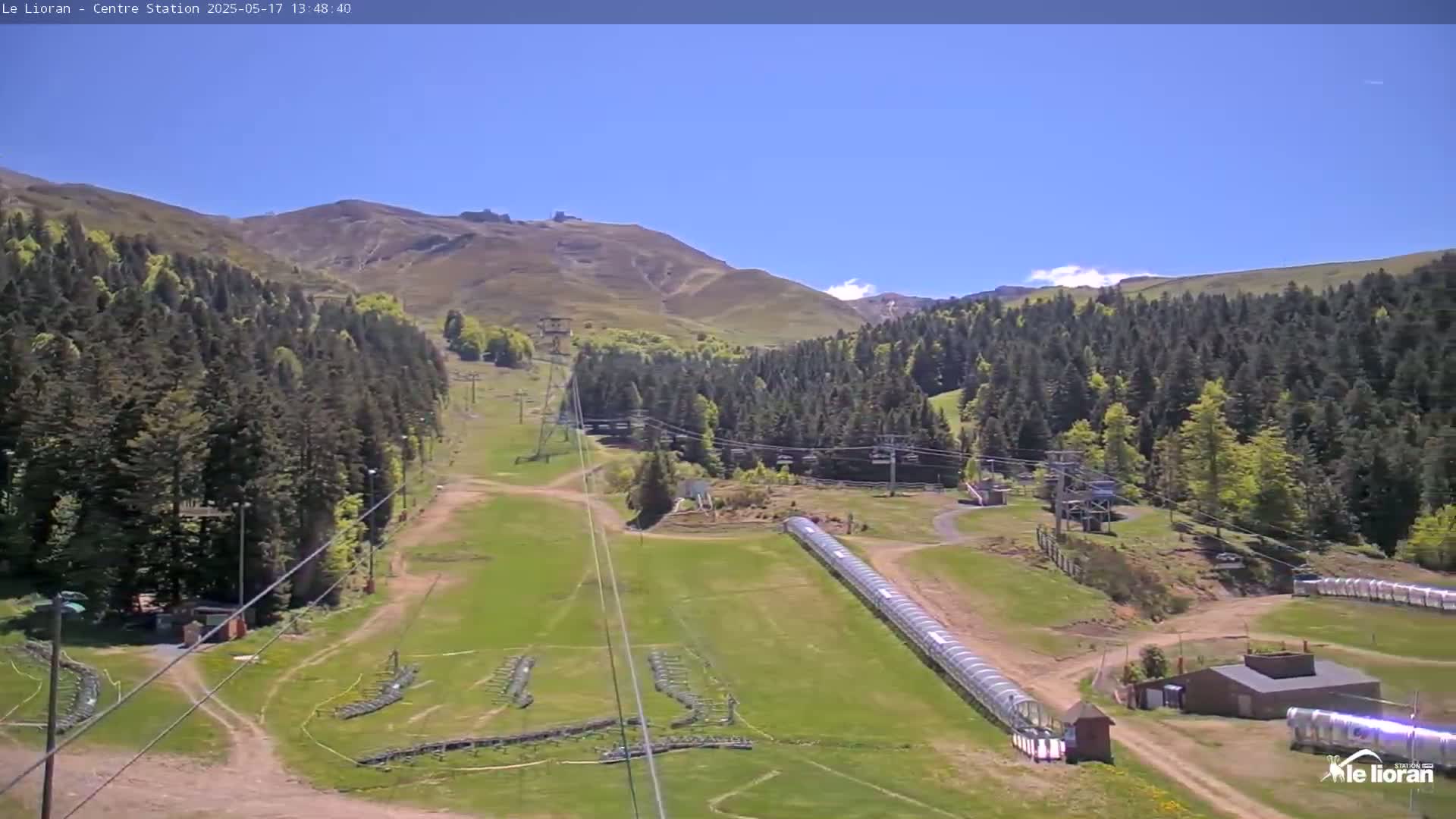 A sunny view of a ski resort in the mountains, showing chairlifts, grassy slopes, and a covered conveyor belt.