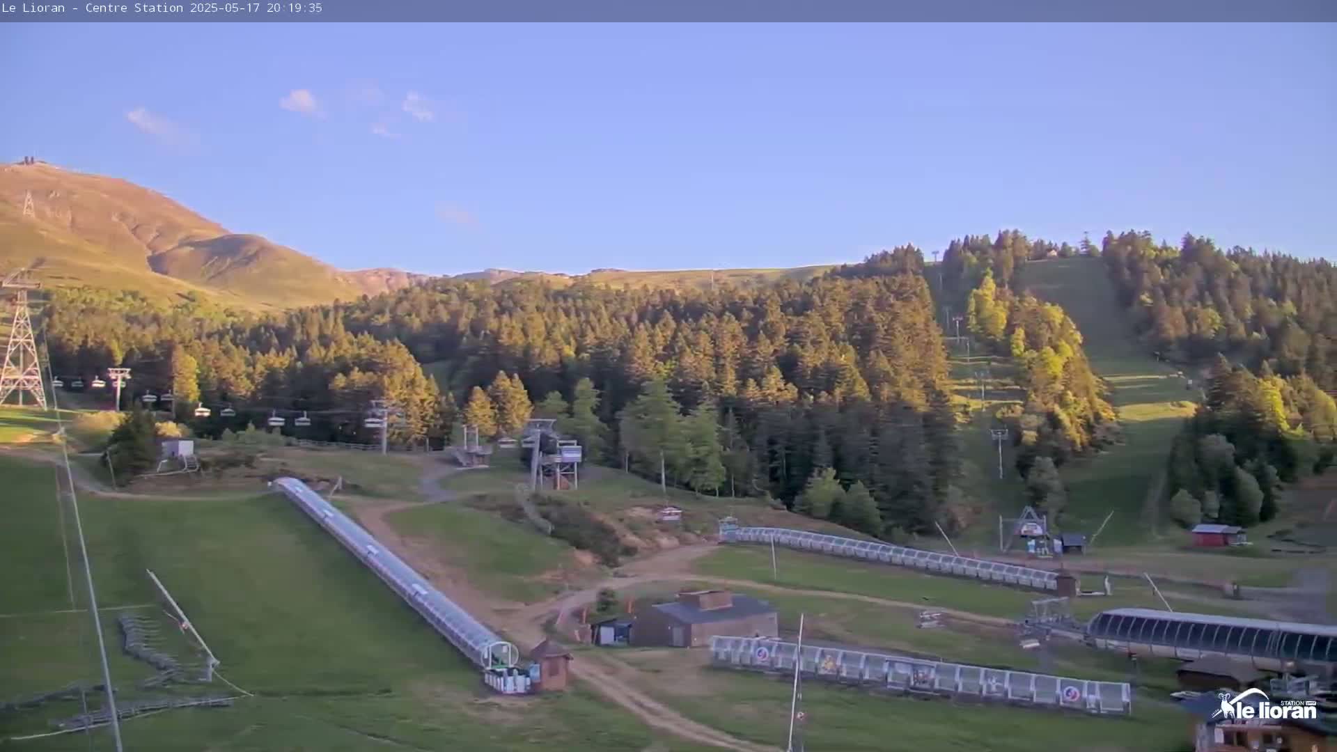 A grassy mountainside ski resort, under a clear blue sky, features chairlifts, a covered ski lift, and several small buildings.