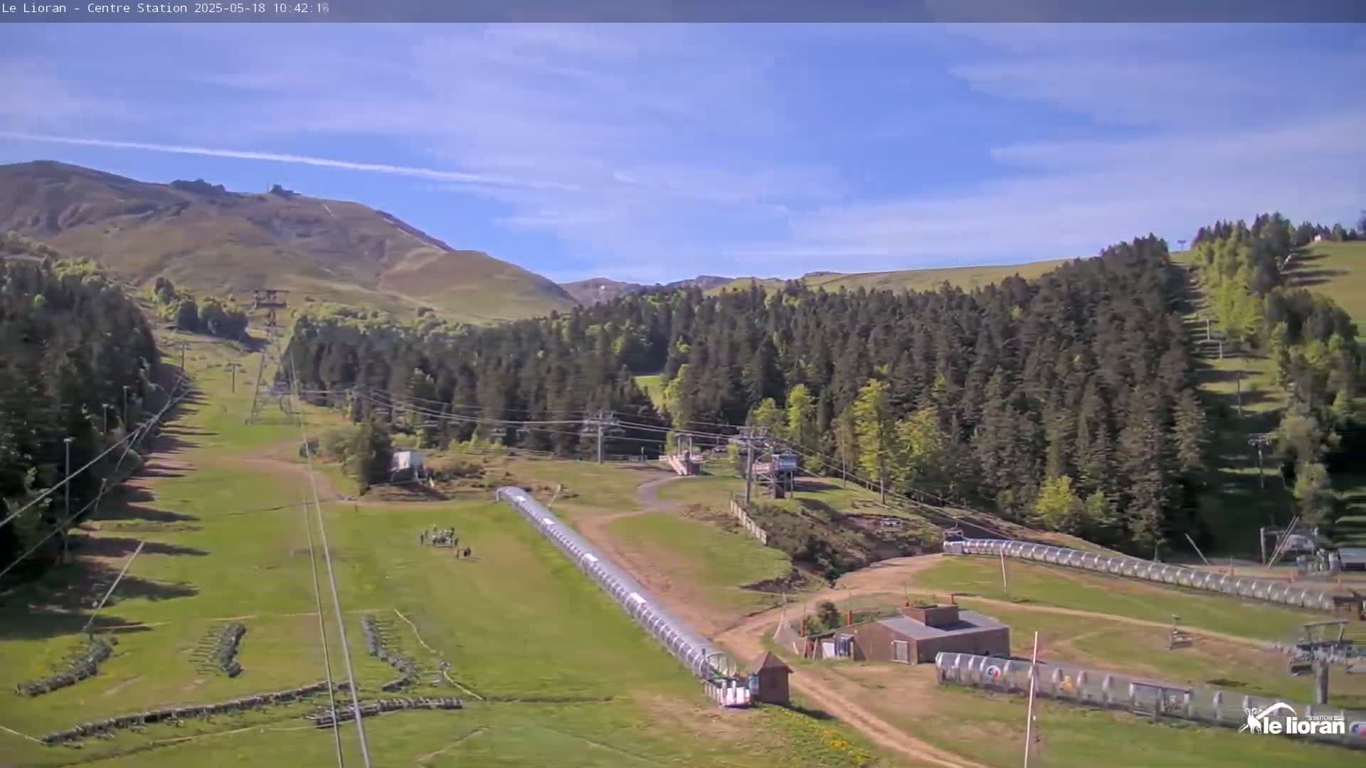 A grassy mountain slope with ski lifts, a long covered structure, and several small buildings under a mostly clear, sunny sky.