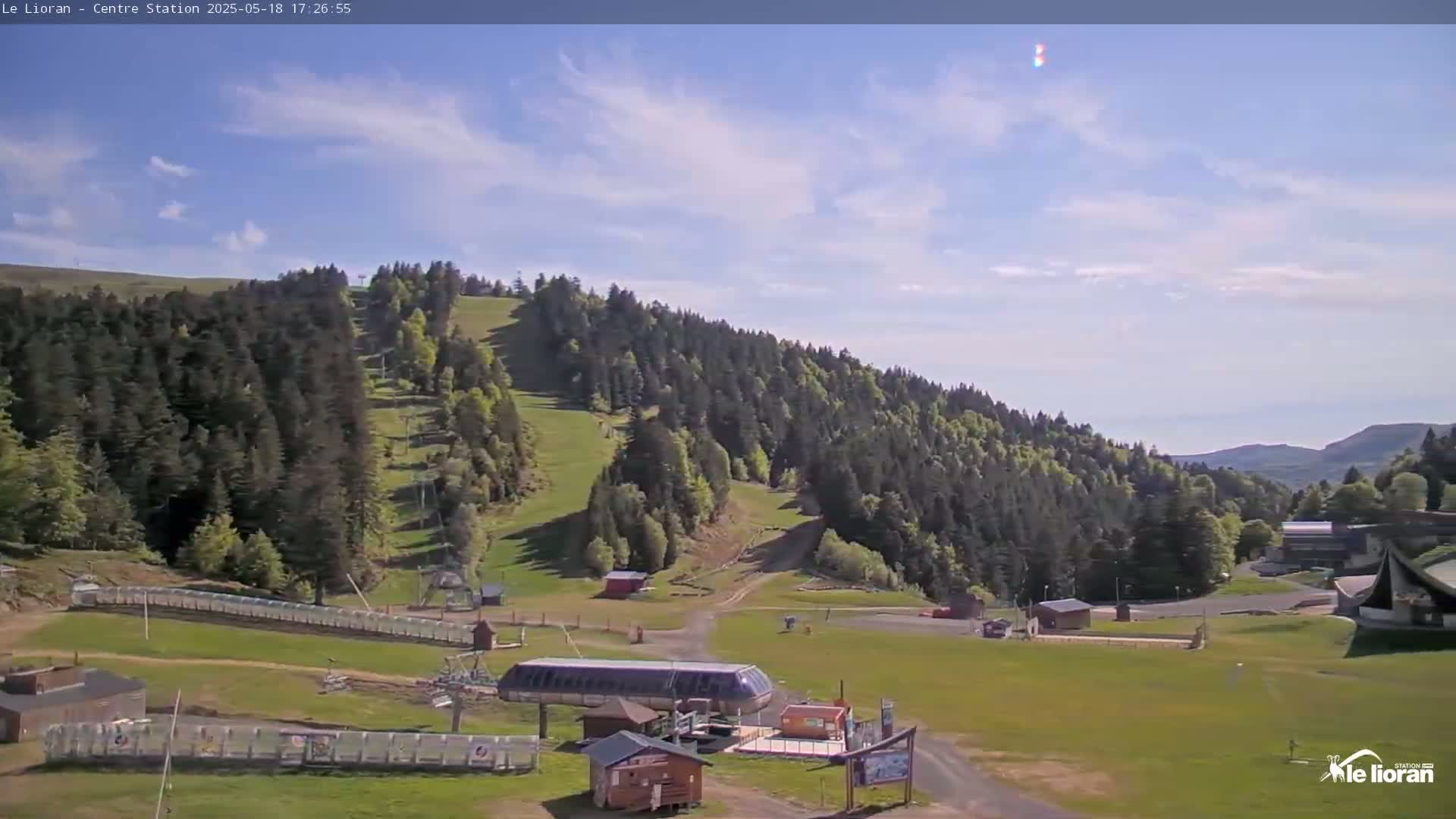 A partly cloudy sky overlooks a grassy mountainside with a ski lift and several buildings, surrounded by evergreen trees.