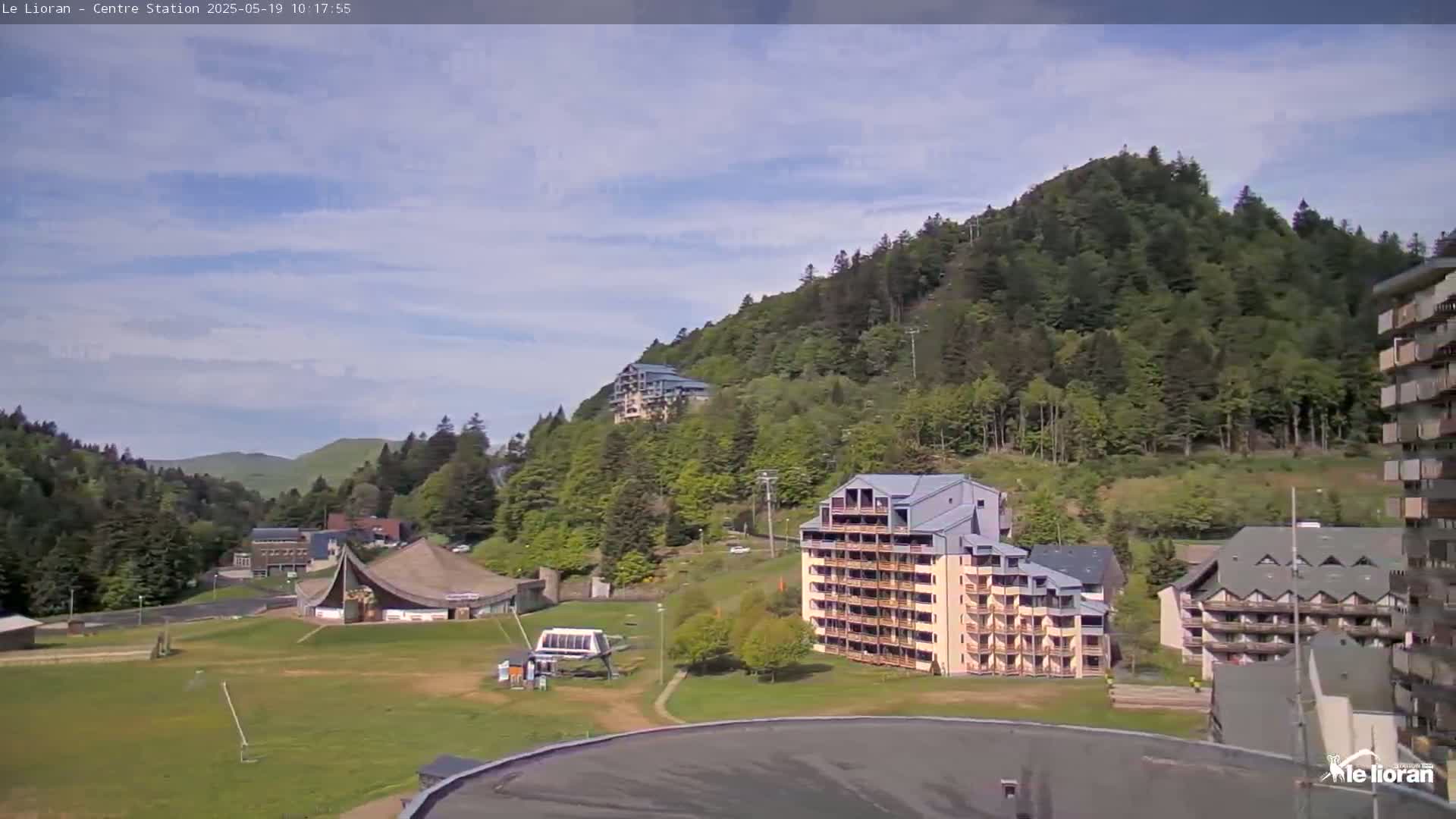 A sunny day reveals a mountainside covered in lush green trees, with several multi-story buildings and a large open grassy area in the foreground.