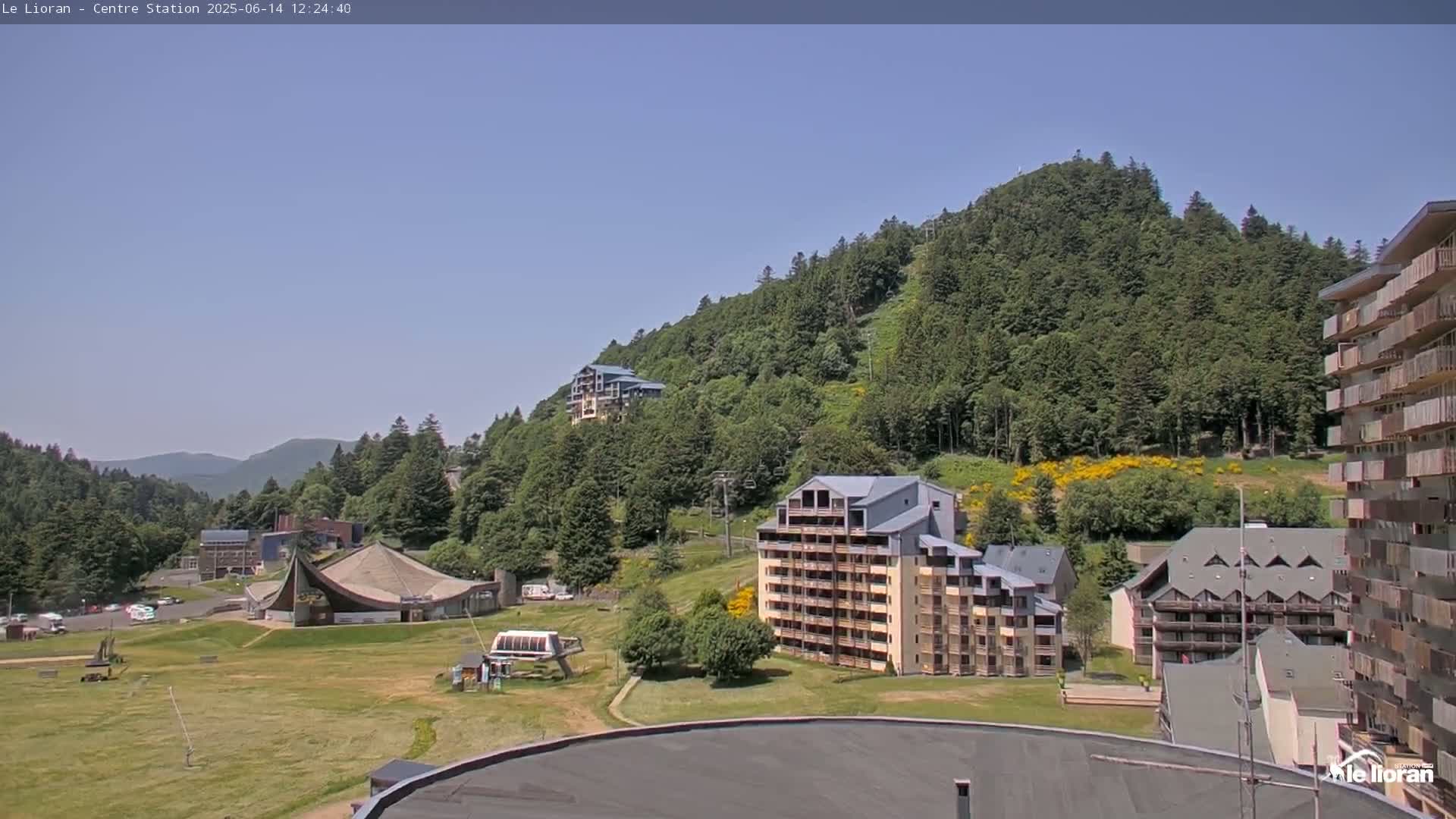 A mountainside covered in lush green trees, with several multi-story buildings and a large grassy area in the foreground, under a clear blue sky.