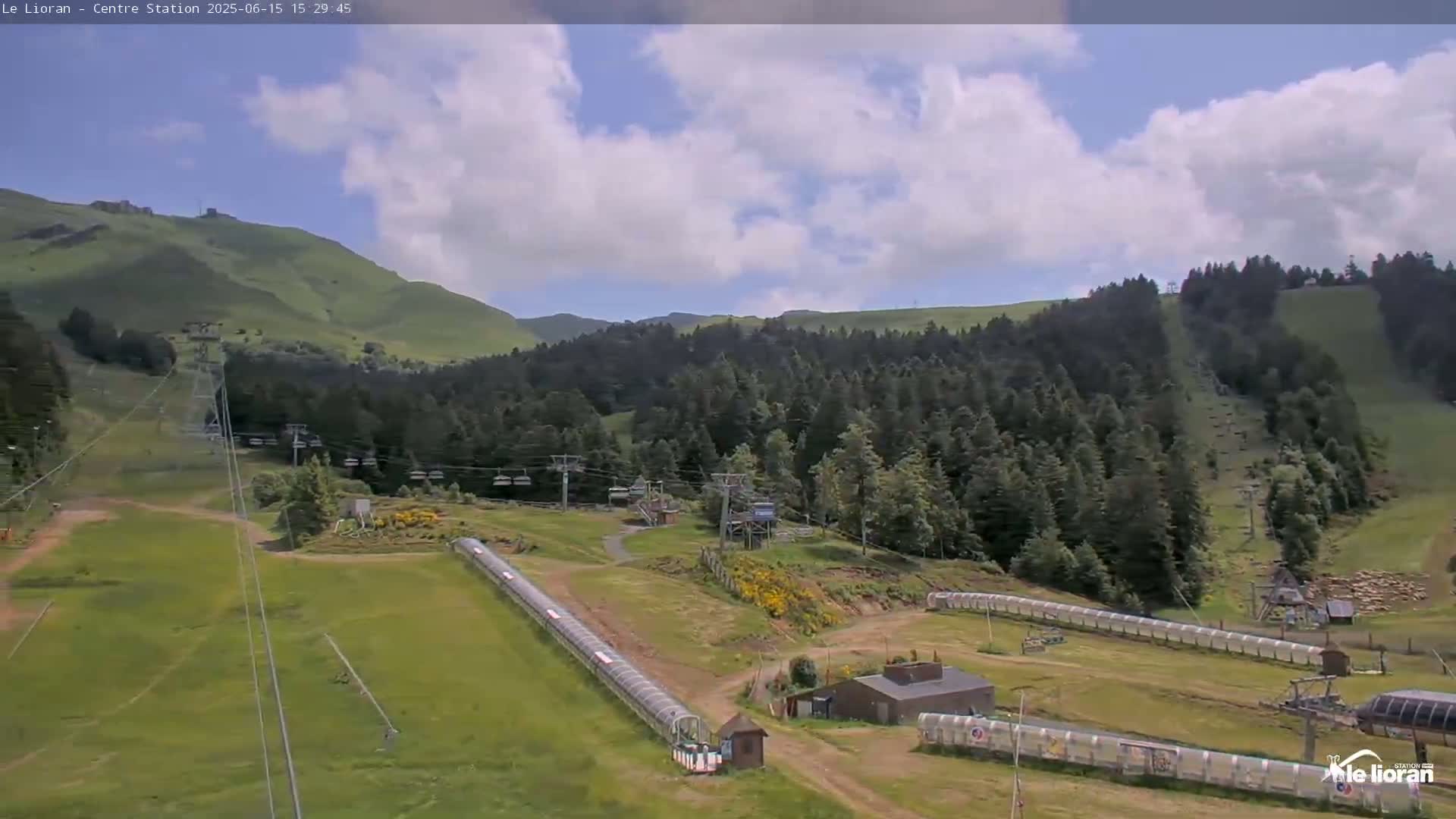 A partly cloudy day reveals a mountainside ski resort with chairlifts and grassy slopes, featuring long, covered structures likely for summer activities.